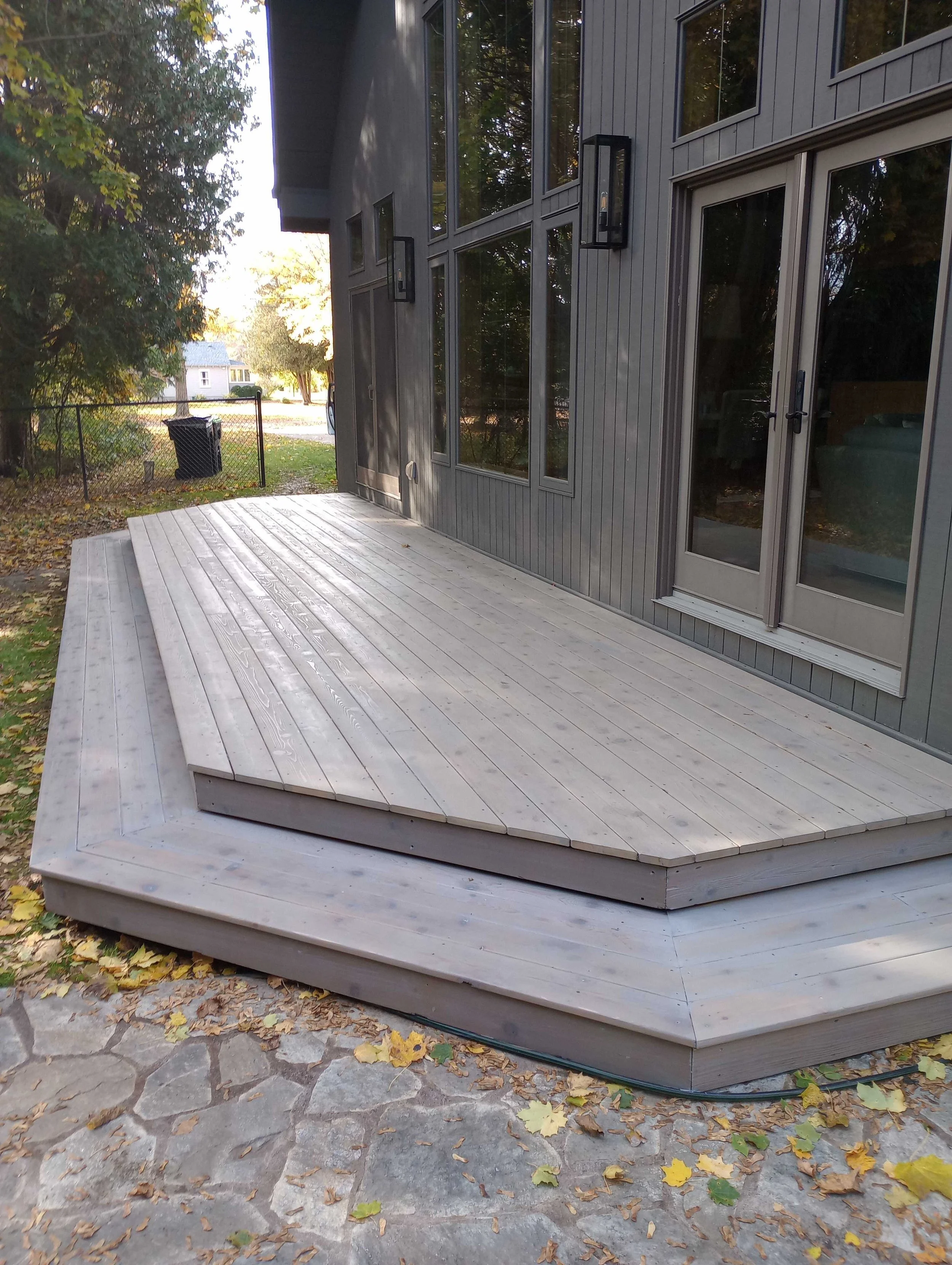 A finished wooden deck with multiple tiers extends from the back of a house with large windows and a sliding glass door. The deck is surrounded by fallen autumn leaves.