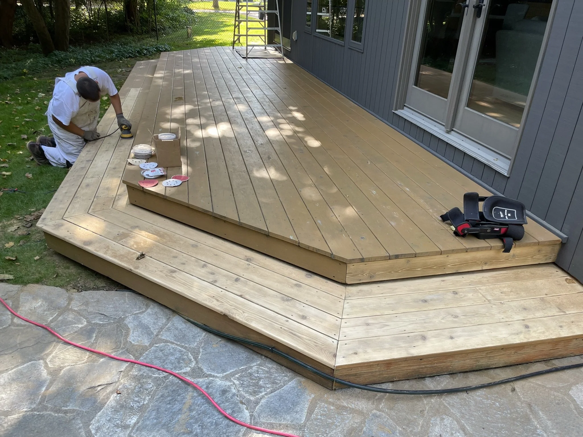 Worker installing wood decking on a porch, kneeling and using a power drill, with tools and materials nearby, adjacent to a house with gray siding and glass sliding doors.