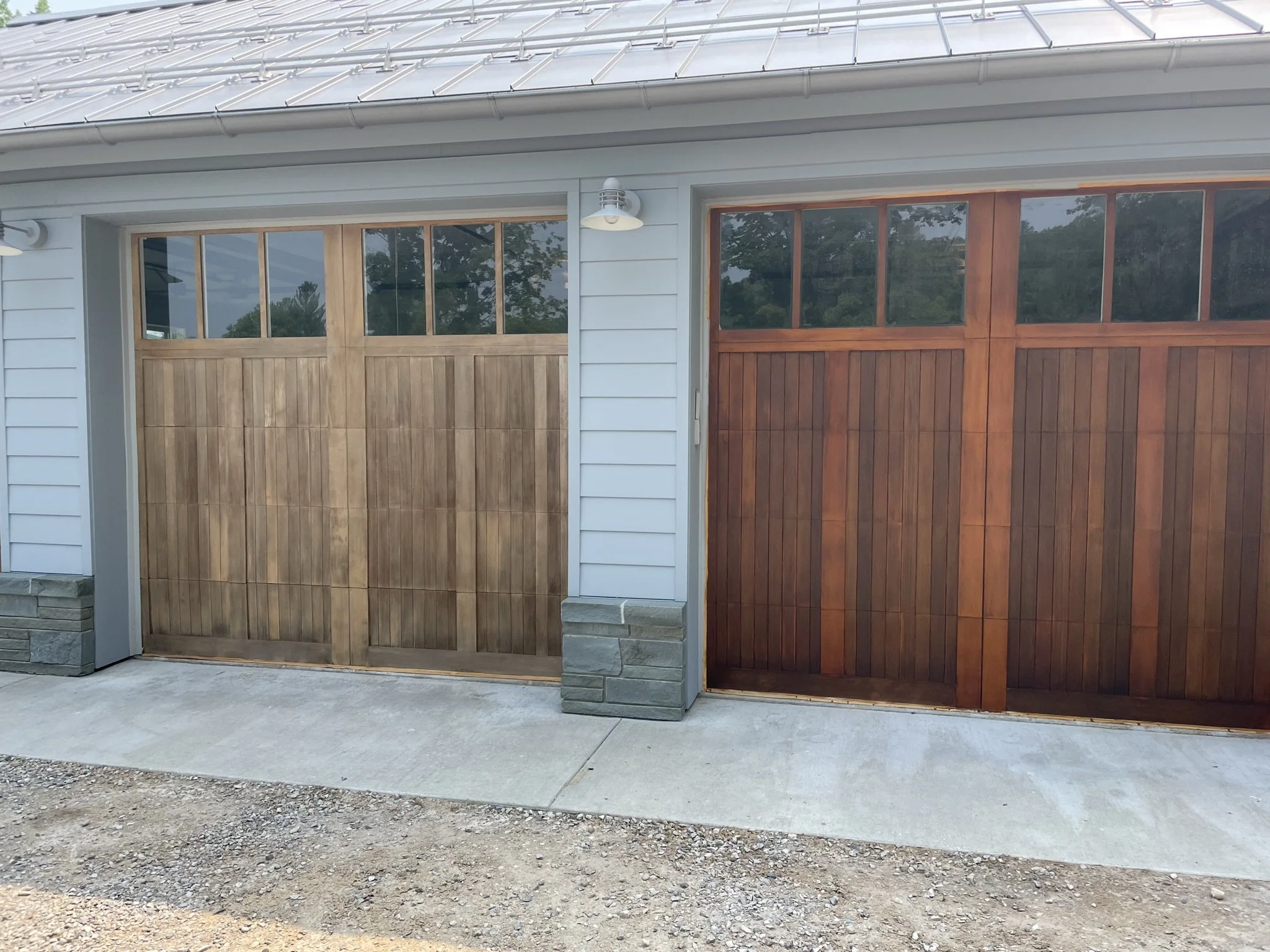 Two wooden garage doors with glass panels at the top, one with a weather worn wood finish and the other with a darker stain finish, set in a light gray house with horizontal siding, stone accents at the base, and exterior lights above.
