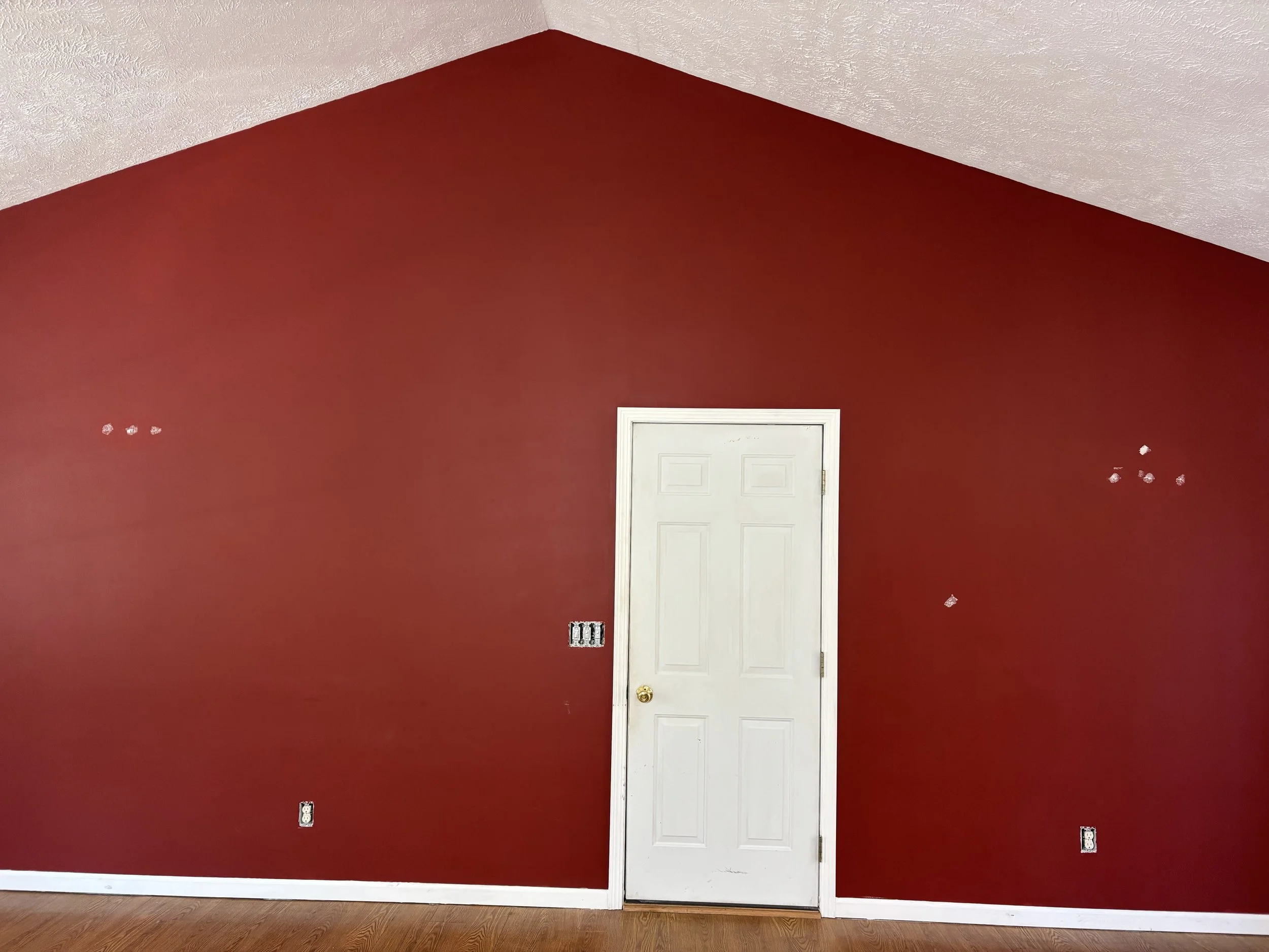 Before picture of an empty red wall with a white door in the center, holes and scratches on the wall, some electrical outlets, and a wooden floor.