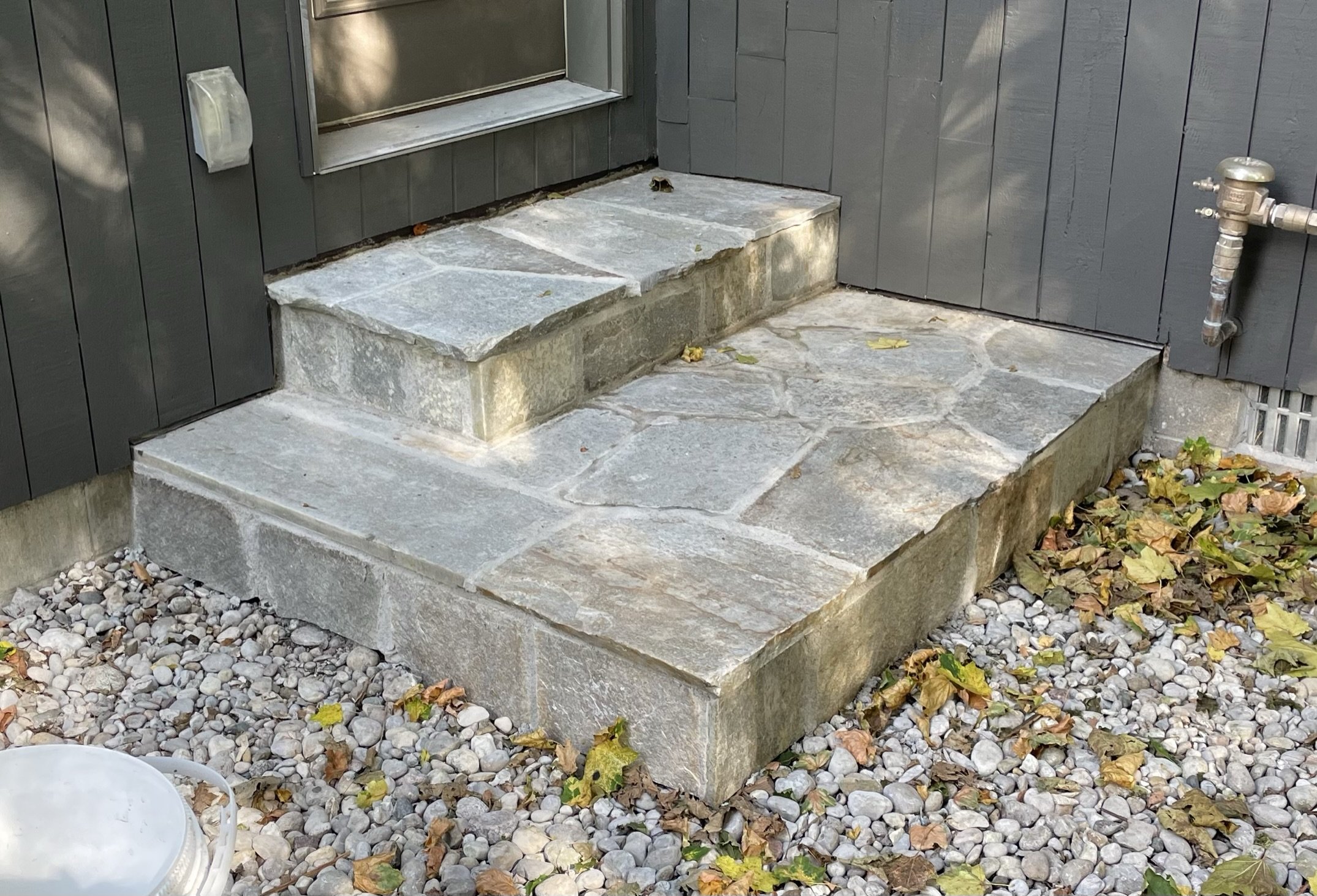Stone steps leading to a door with dark gray siding, surrounded by small gravel and fallen leaves.