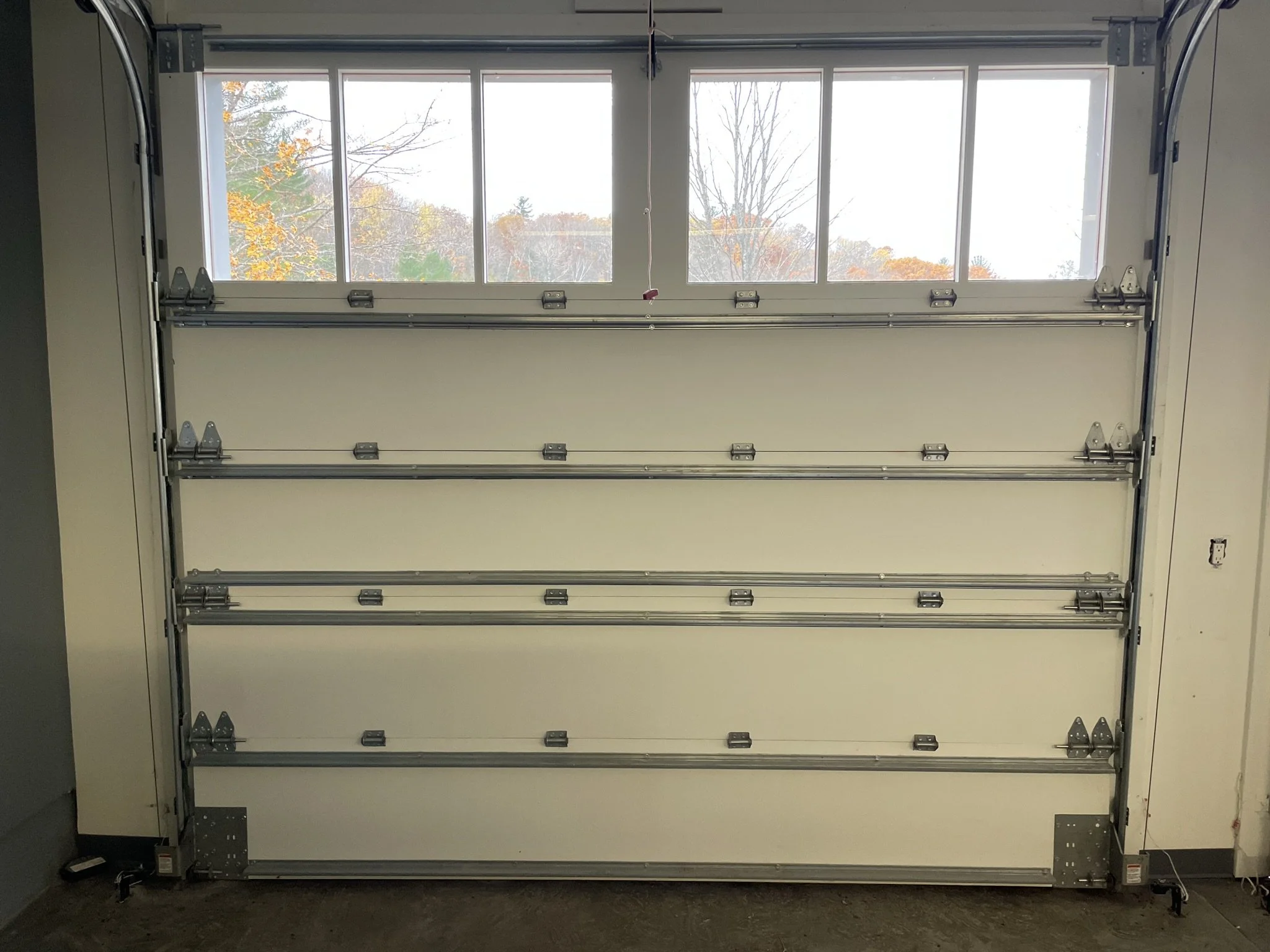 A white garage door with four horizontal panels, each with metal tracks and hinges, and a window at the top revealing an autumn landscape with trees and colorful leaves. Before.