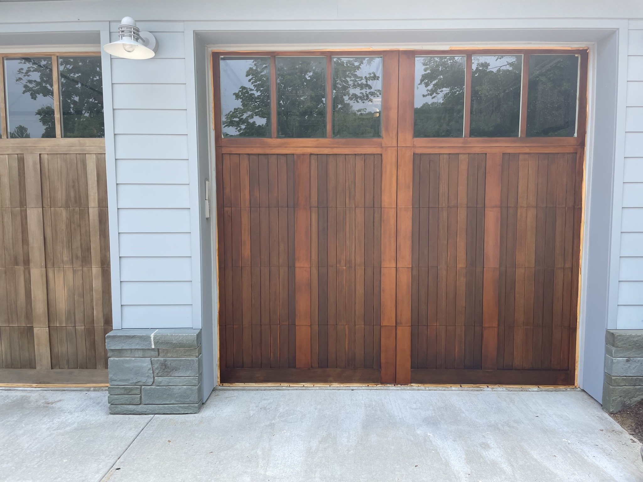 A finish stained wooden garage door with four glass panes at the top, framed by white siding and gray stone accents at the bottom corners, and a modern exterior light fixture on the left.