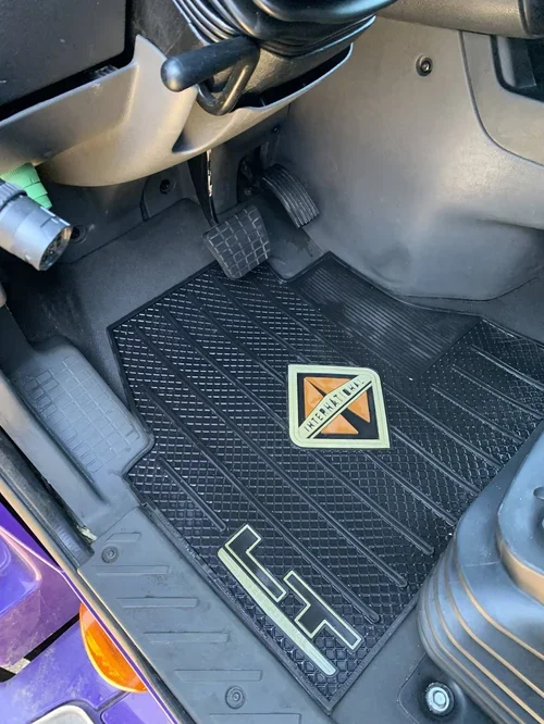 The driver’s side floor of a vehicle with a black rubber mat, a warning sticker, and a few stickers on the mat.