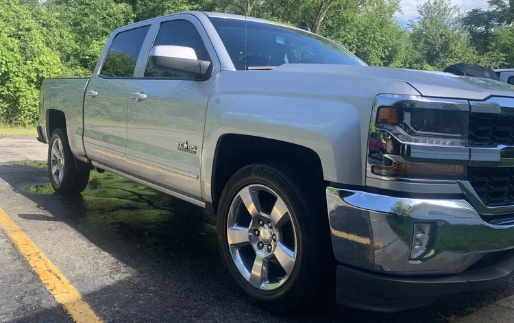 Front and side view of a silver pickup truck parked on a paved surface with green trees in the background and a yellow parking line in the foreground.