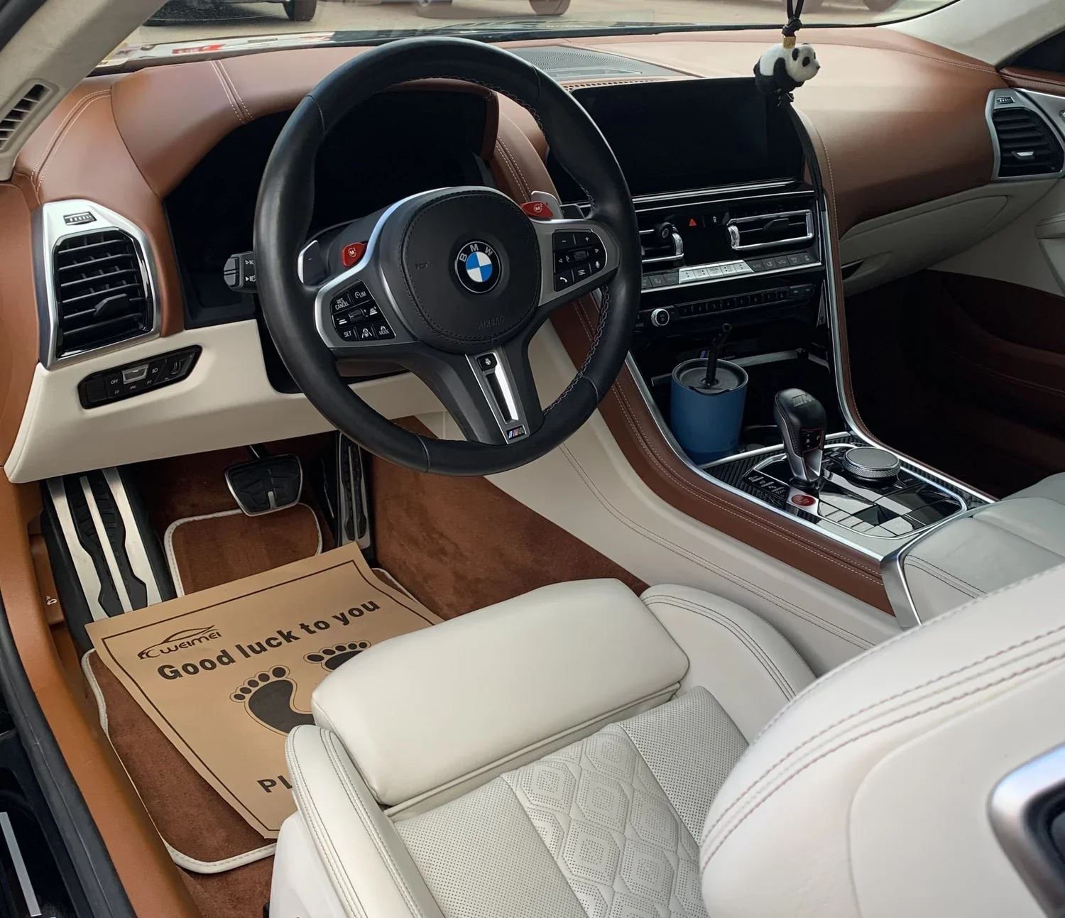 Interior of a luxury BMW car with tan and white seats, a tan dashboard, and a black steering wheel, featuring a gear shift, cup holder with a blue drink, and a panda plush hanging from the rearview mirror.