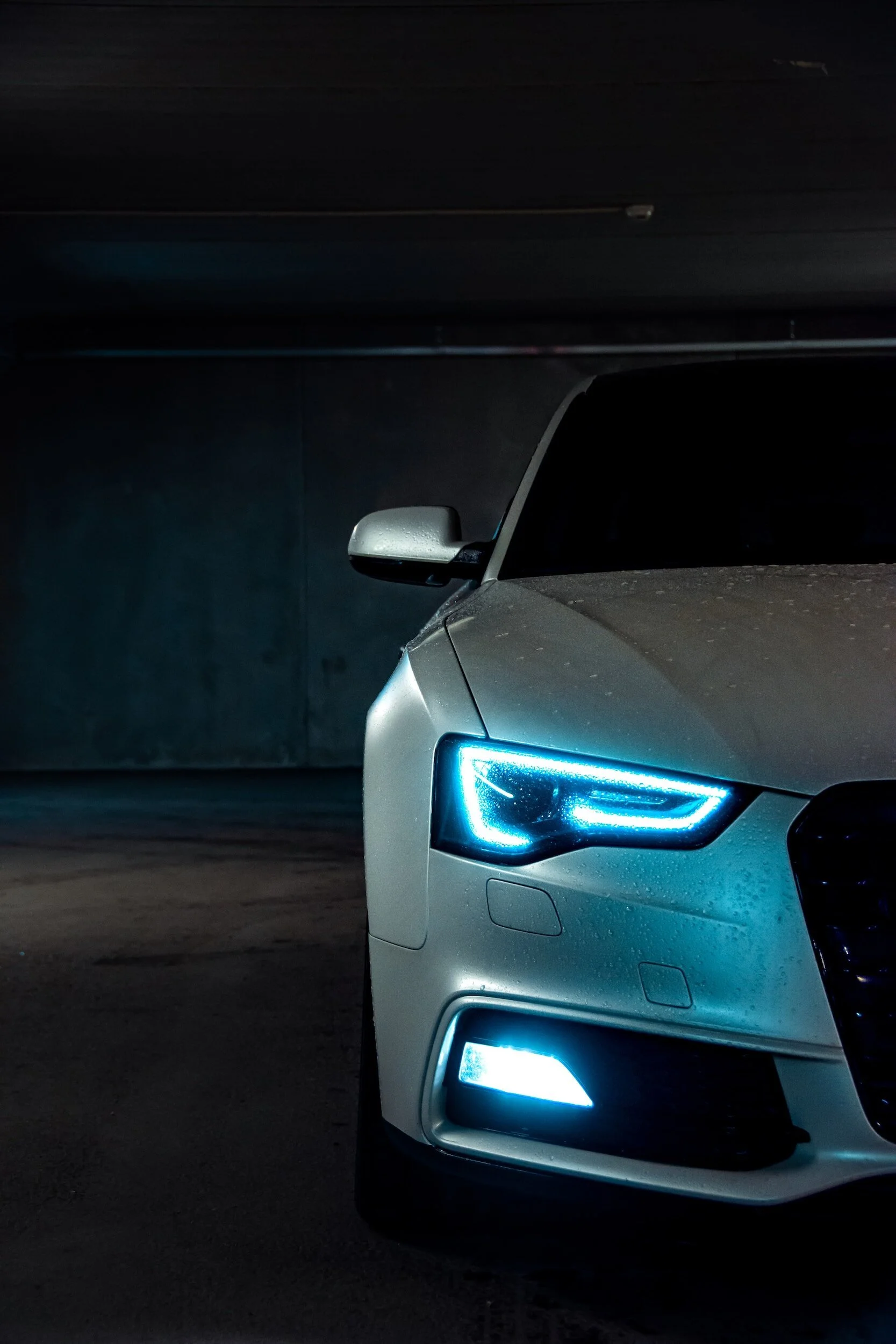 Close-up of the front right side of a white car with illuminated blue headlights, parked in a dark parking garage.