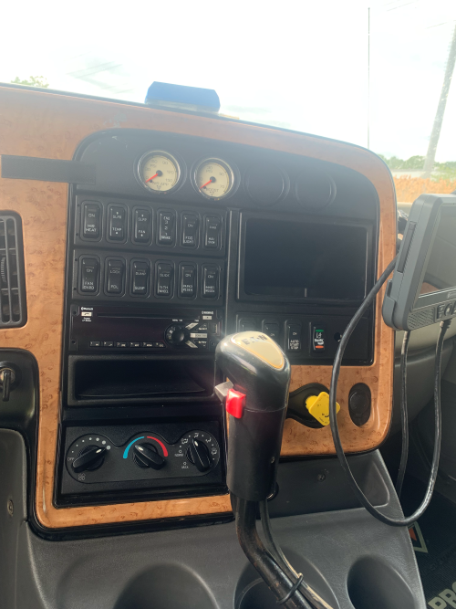 Dashboard of a truck cab with gauges, control buttons, a display screen, and a gear shift lever, with a mobile device mounted on the right.
