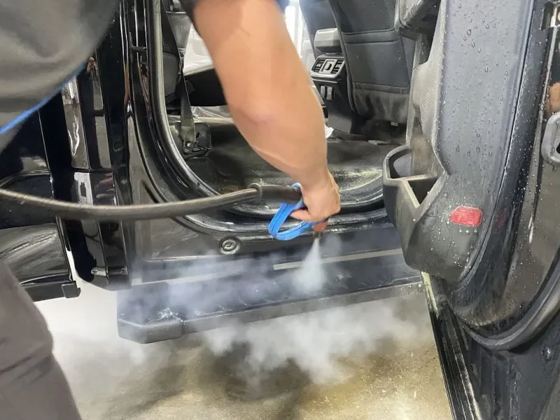 Person using a spray tool to clean the interior of a vehicle, with visible steam or spray coming from the tool.
