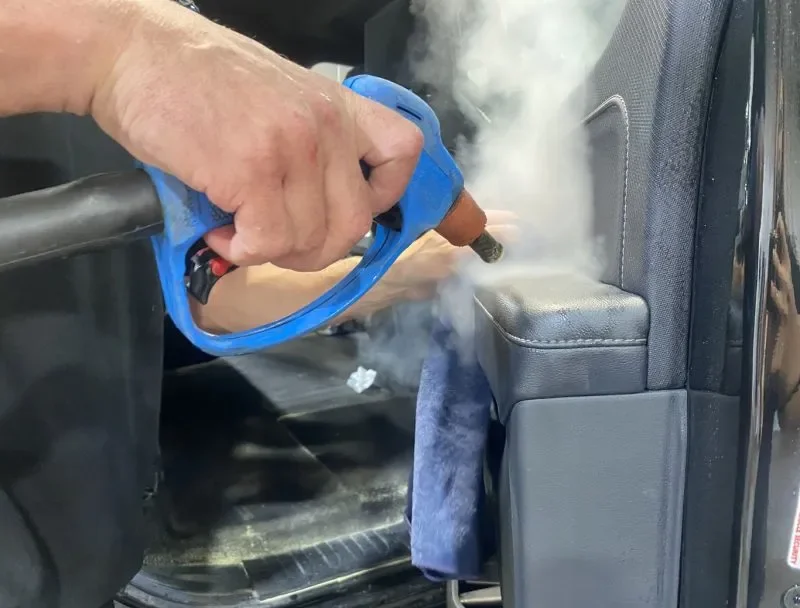 A person is using a handheld steam cleaner to clean the interior of a vehicle, applying steam to the seat area.