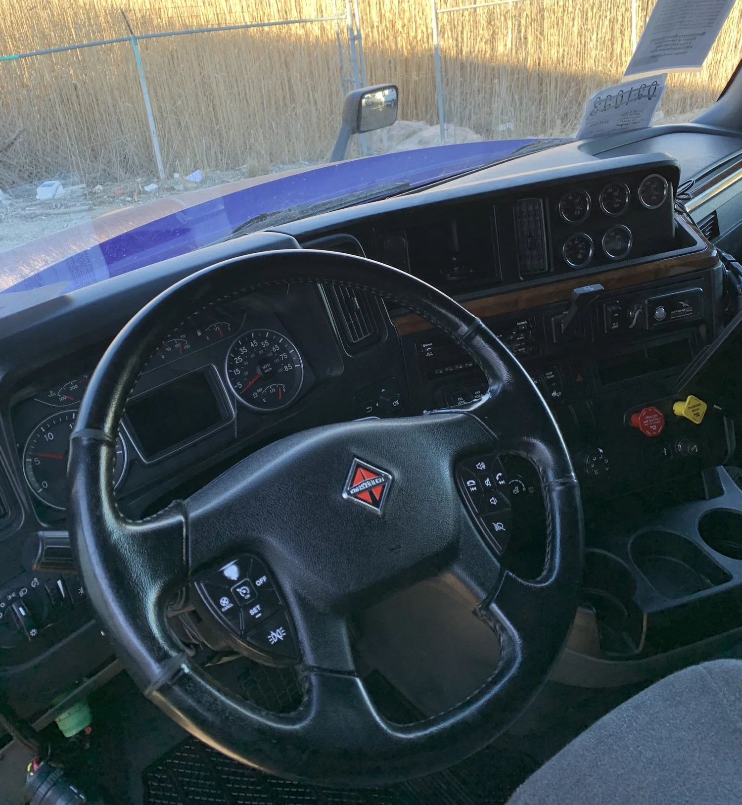 Interior of a truck cab, showing a steering wheel with a Hazardous Materials sign, dashboard with gauges, and cup holders.