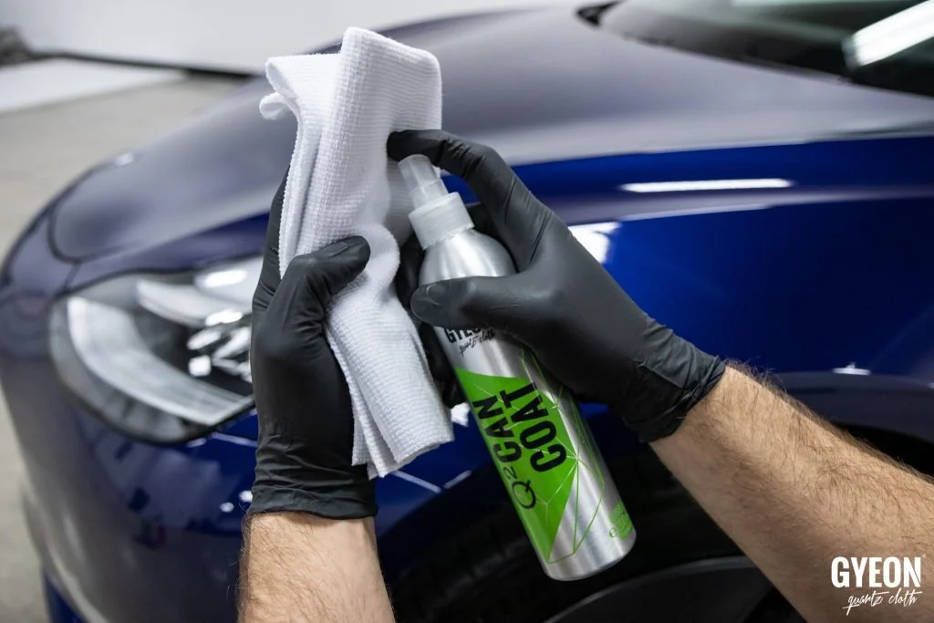 Person in black gloves cleaning a car's blue surface with cleaning spray and cloth.