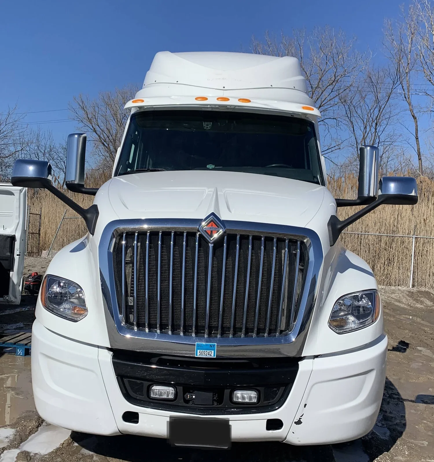 Front view of a white semi-truck with large chrome grille, black accents, and side mirrors, parked outdoors on a sunny day with trees in the background.