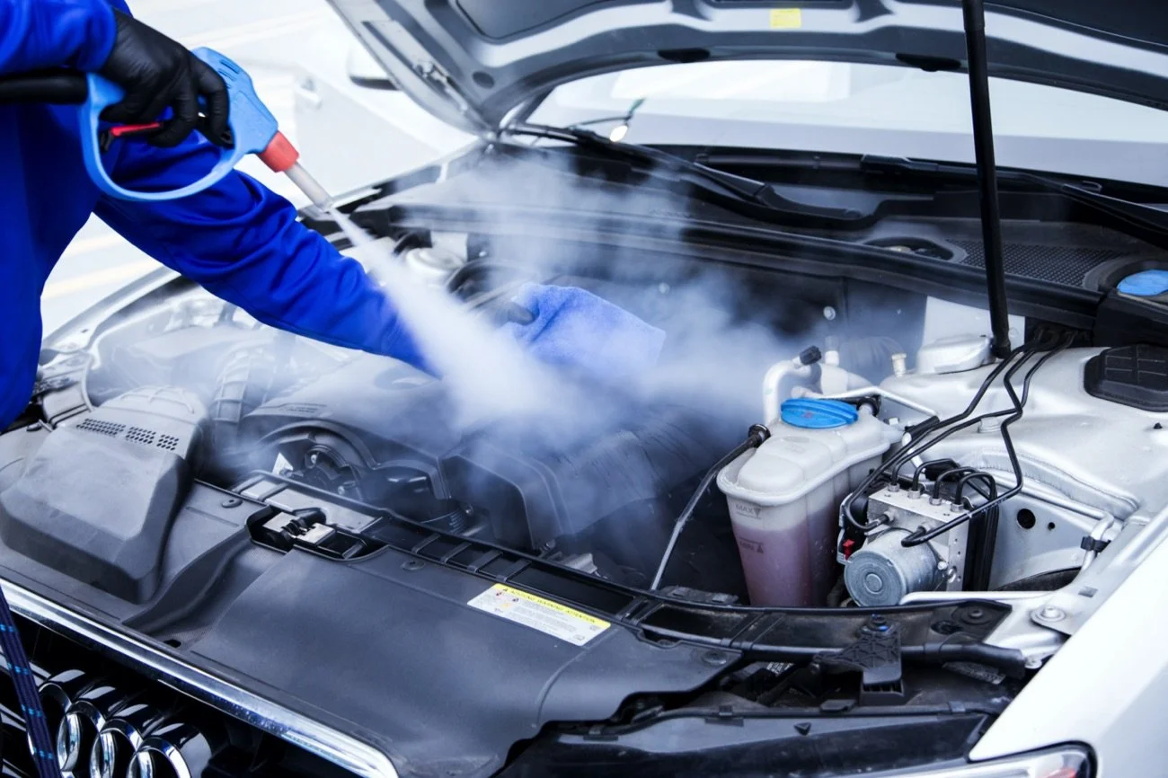 A mechanic wearing gloves sprays a cleaning substance into the engine bay of a car, emitting a cloud of cleaning mist.