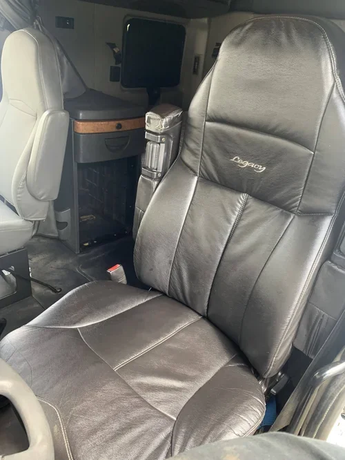 Inside an airplane cabin, showing a leather passenger seat labeled 'Legacy' with headrest, next to another seat and a small table or console.