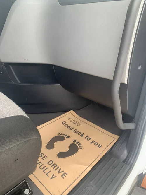 Car floor with a paper floor mat that has footprints and the message 'Good luck to you. Please drive safely fully.'