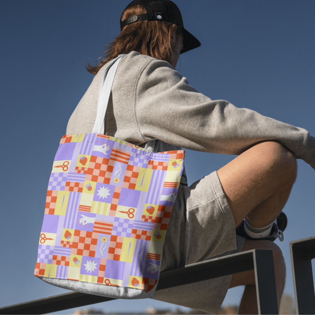 Person sitting on a ledge wearing a gray sweatshirt, beige shorts, a black cap, and white sneakers, with a colorful patterned tote bag hanging from their shoulder, against a clear blue sky.