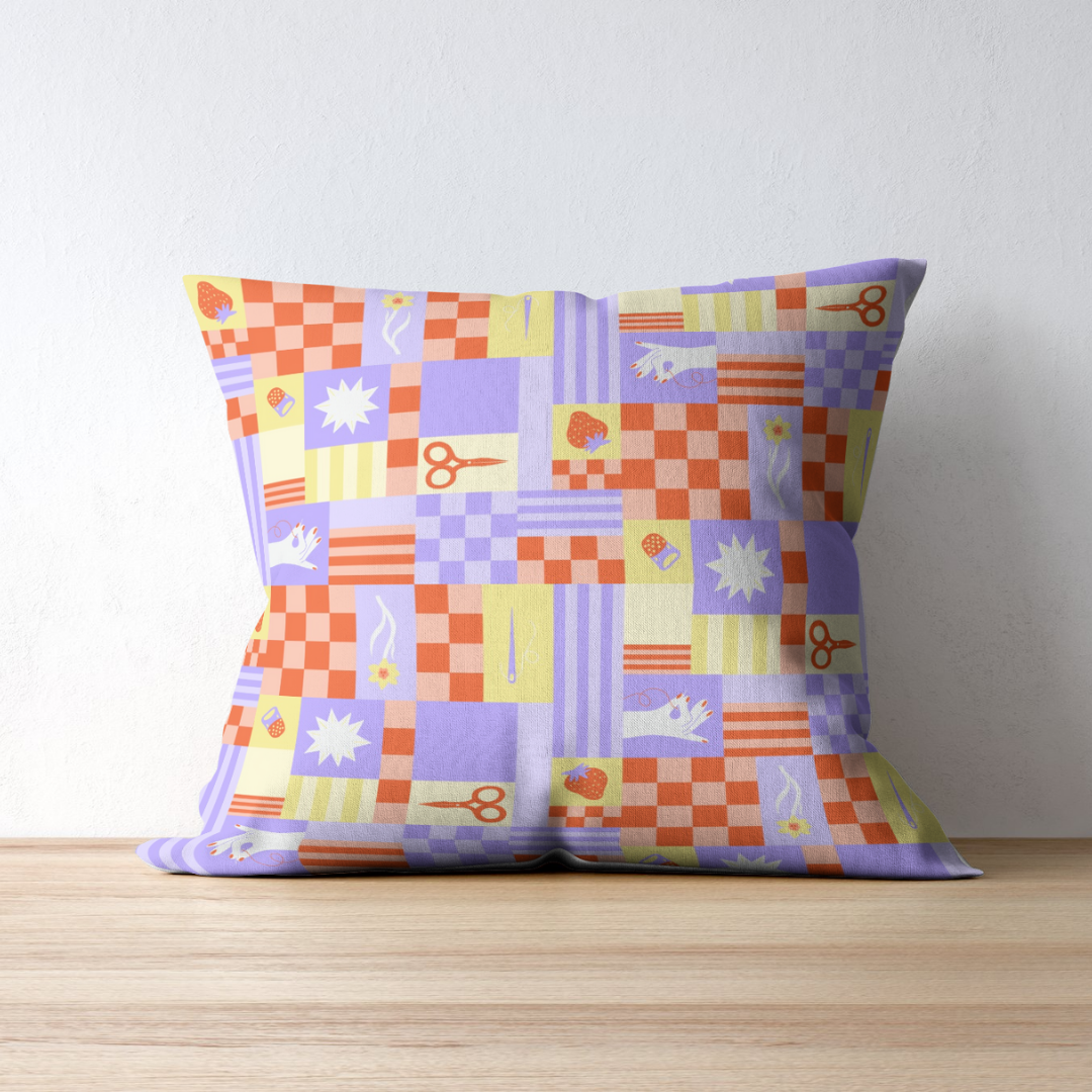 A decorative pillow with a patchwork pattern of various squares in purple, orange, yellow, and lavender, featuring icons like scissors, sewing needles, cupcakes, strawberries, flowers, and stars, placed on a wooden surface against a white wall.