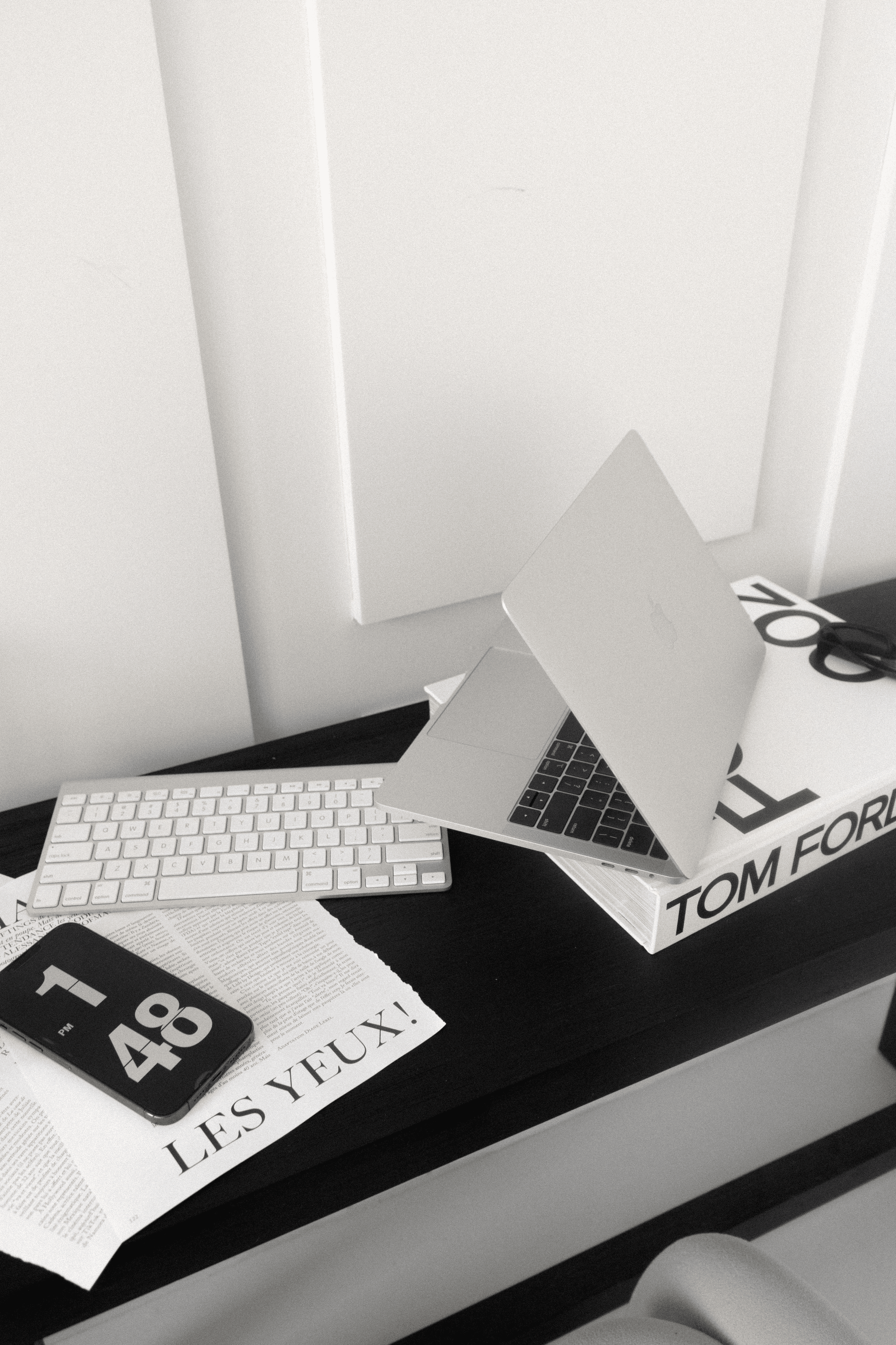 Desk with laptop, wireless keyboard, smartphone, newspaper, and design book in black and white.