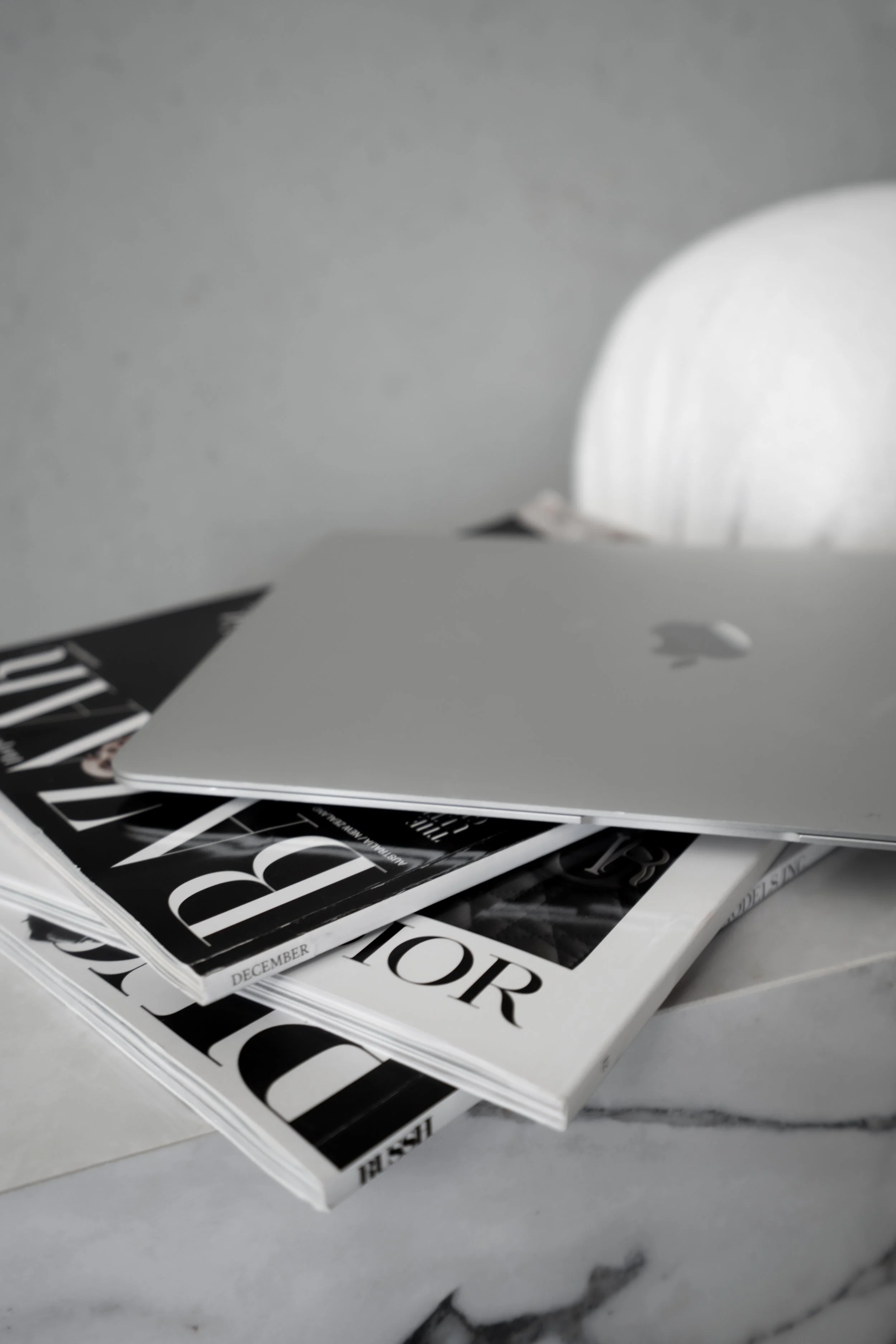 A silver Apple MacBook placed on top of a stack of black and white magazines, with a blurred white object in the background.