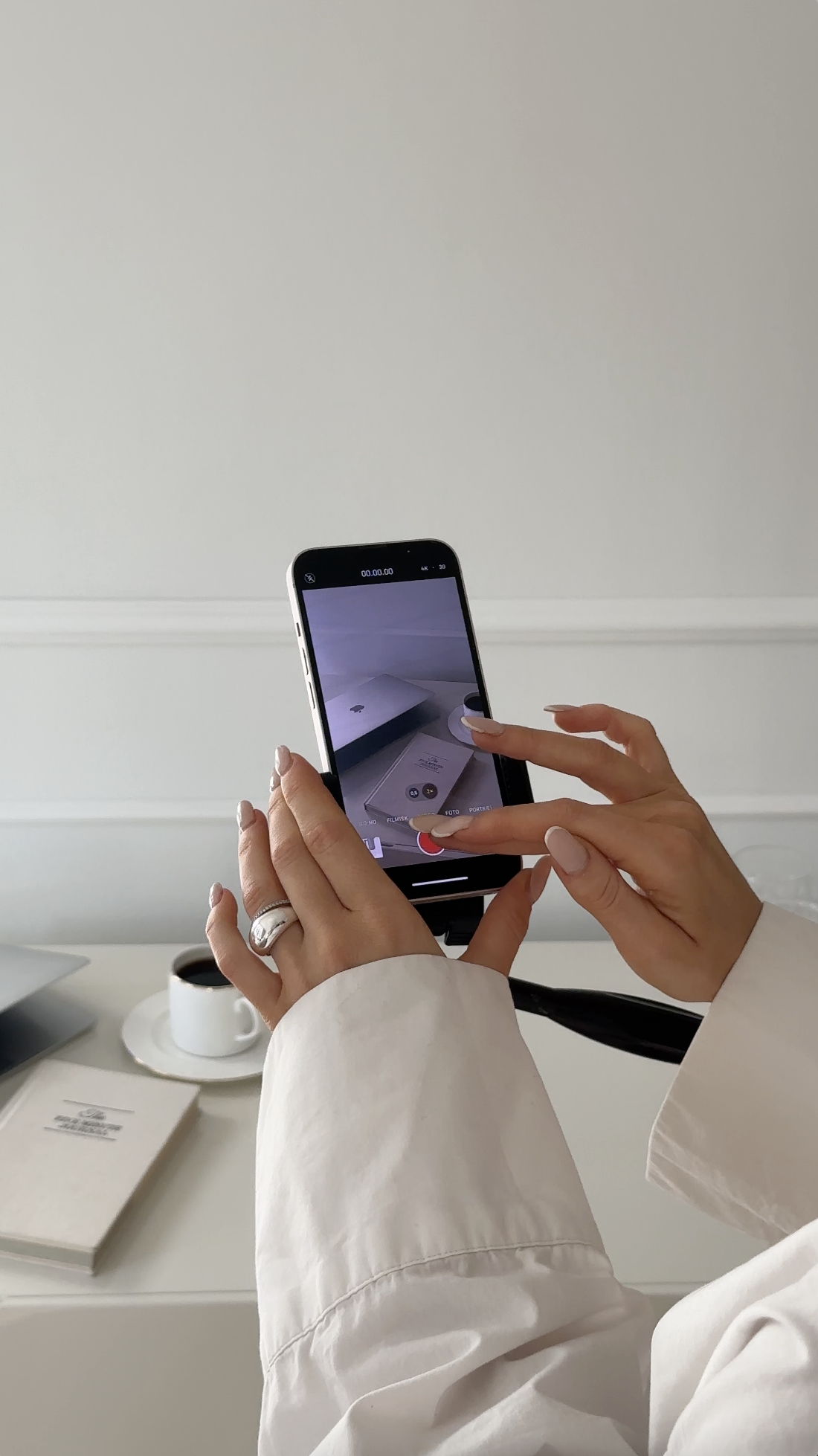 Person taking a photo or video with a smartphone of a white book and a cup of coffee on a table.