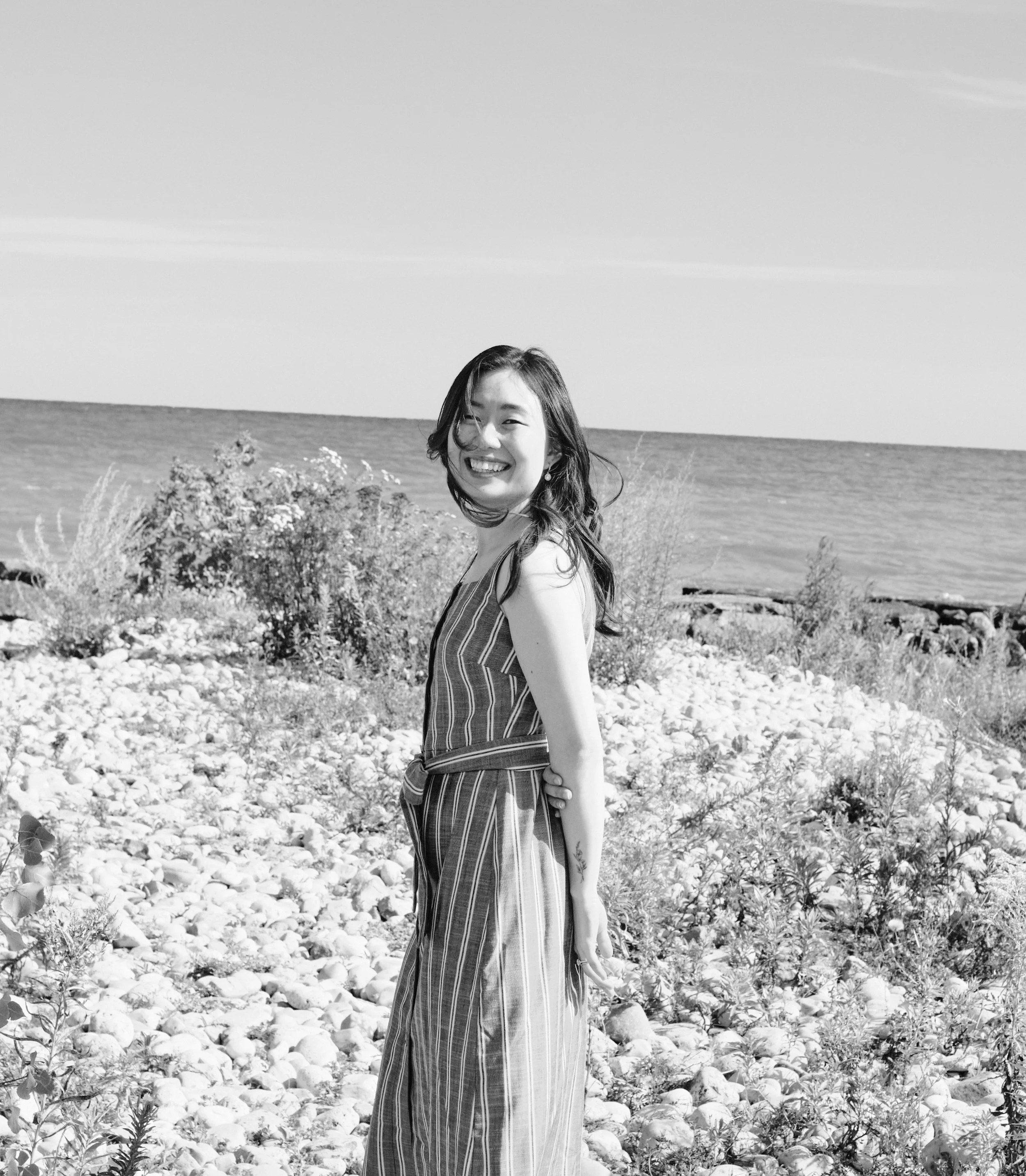 A woman standing on a rocky beach by the ocean, smiling and looking at the camera, in a sleeveless striped dress.