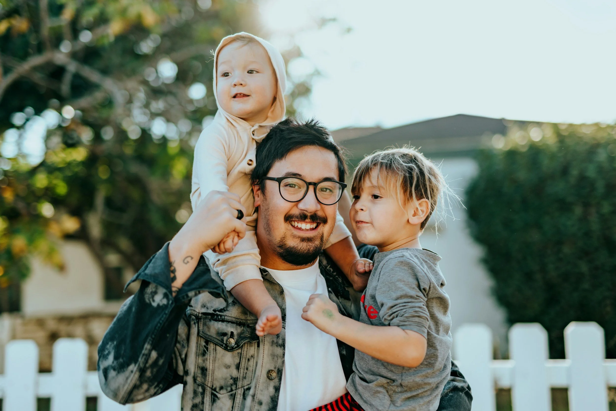 A man with glasses smiling, carrying a young girl on his shoulder and holding a boy. The boy is leaning against his chest. They are outdoors with green trees and a white picket fence in the background.