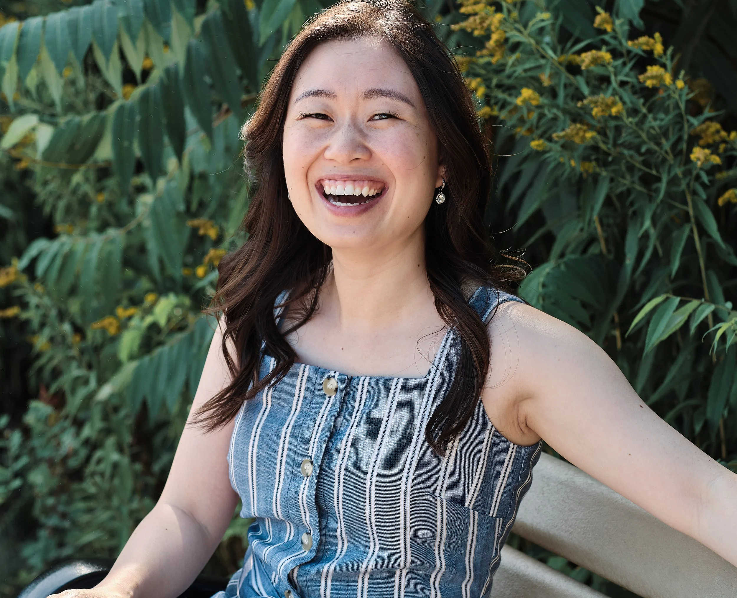 A woman with long dark hair smiling, sitting on a bench outdoors with green foliage and yellow flowers in the background.
