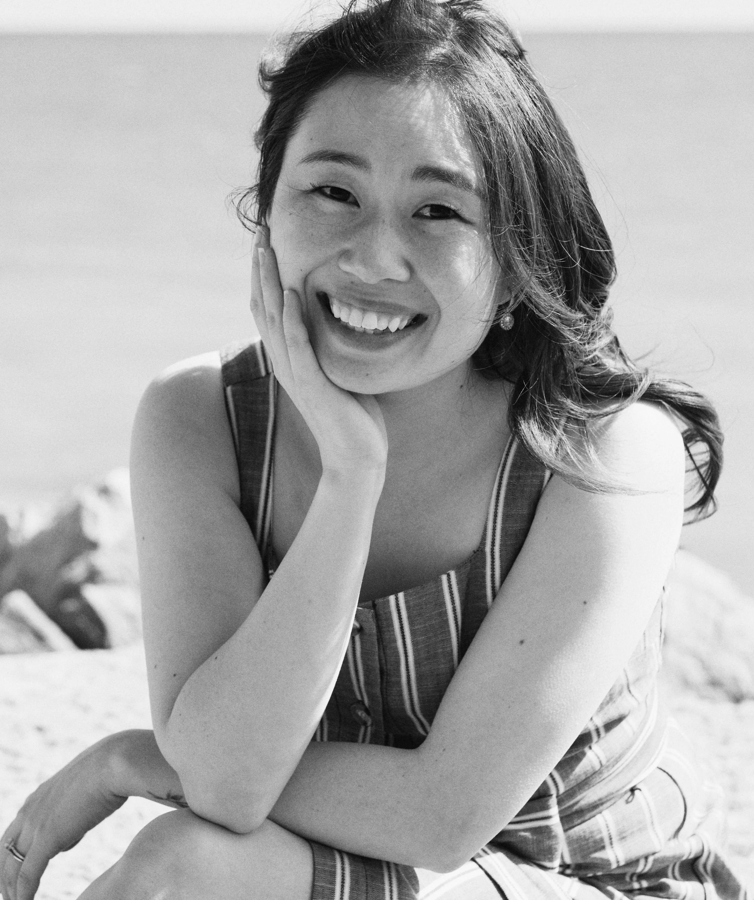A smiling woman with wavy hair in a striped dress sitting outdoors, resting her head on her hand.