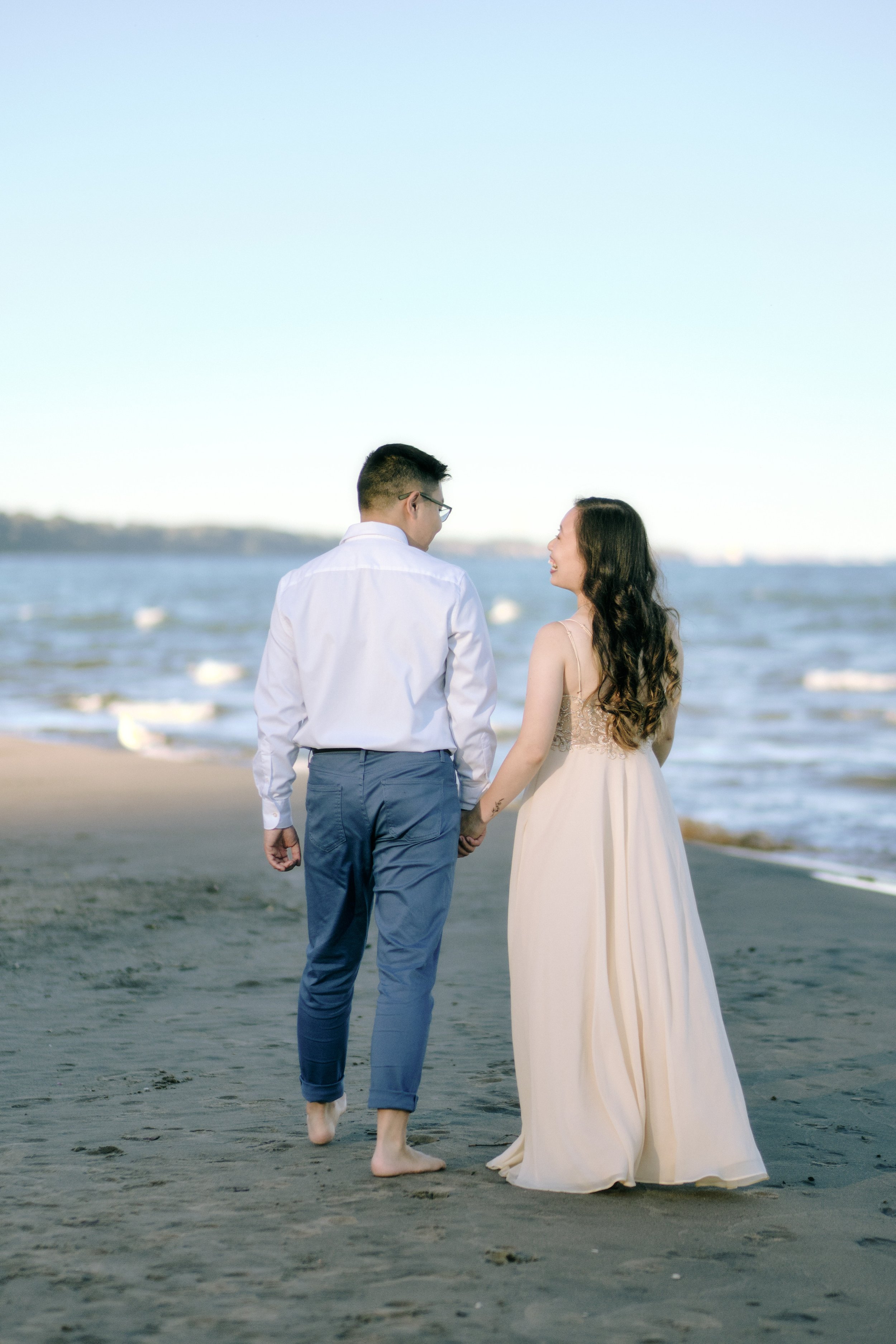 A couple holding hands and walking barefoot on the beach, gazing at each other, with ocean waves in the background.