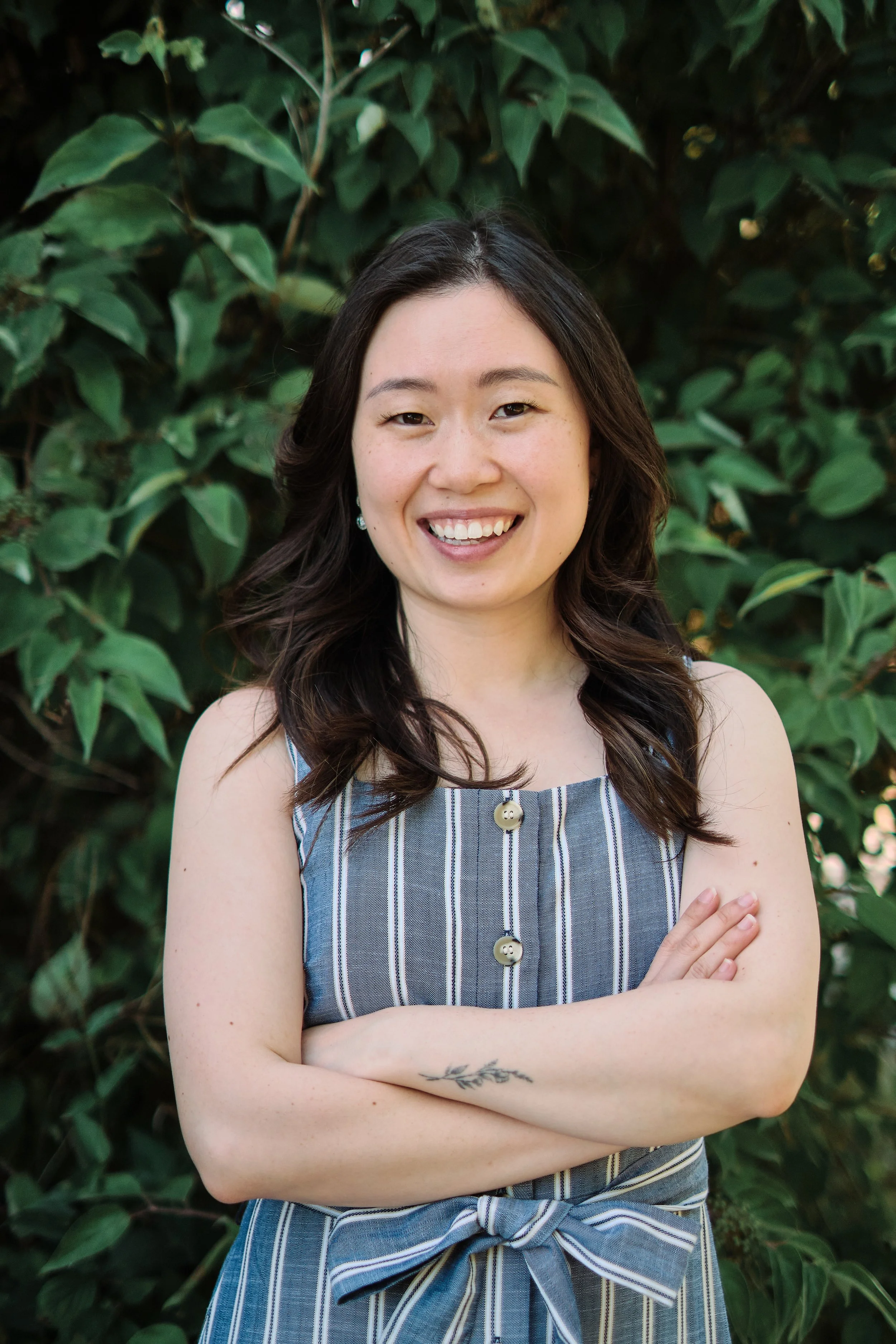 A woman with dark wavy hair smiling and crossing her arms, standing outdoors in front of green leafy bushes, wearing a sleeveless striped dress.