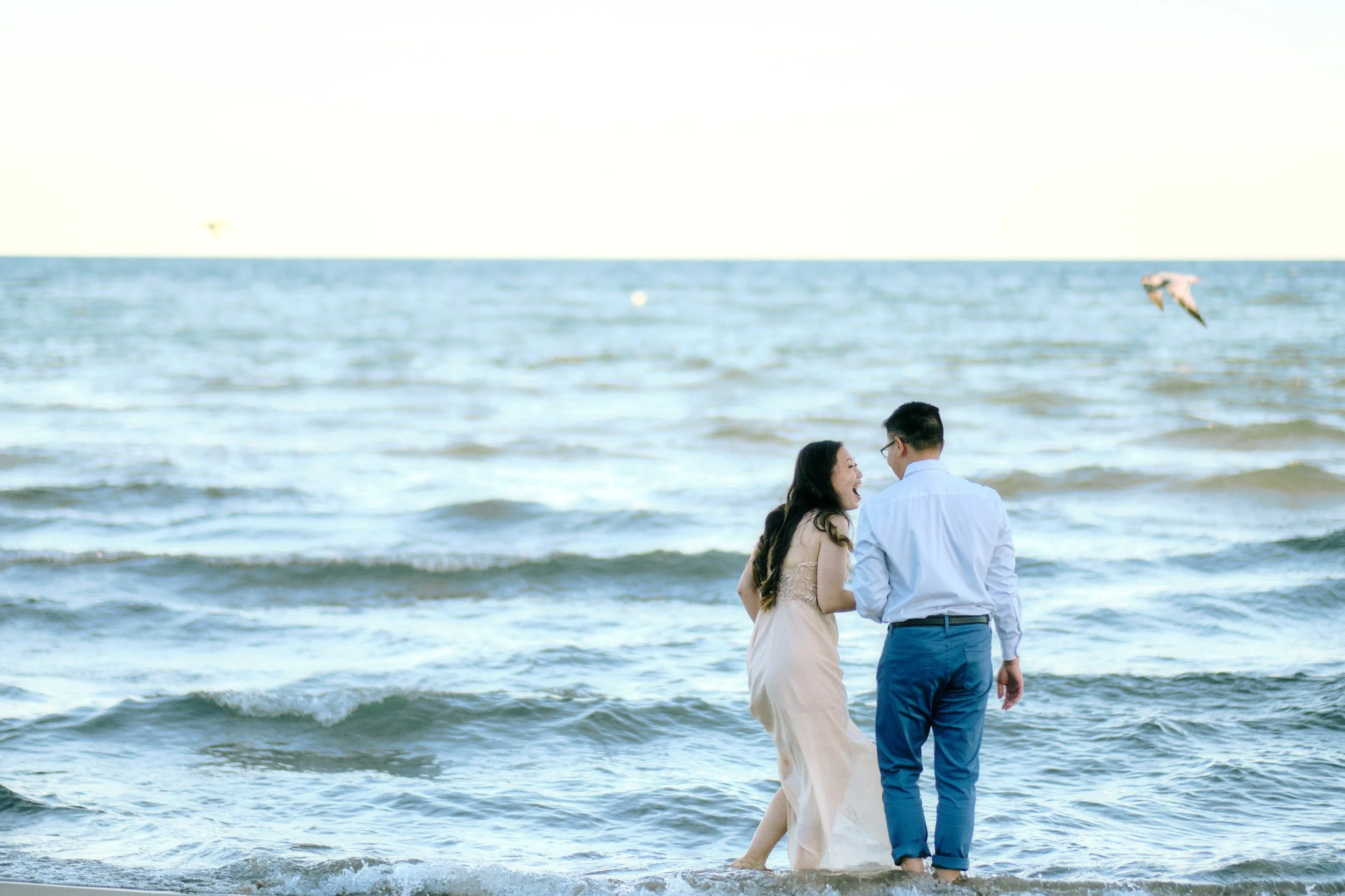 A couple standing in the ocean waves, holding hands, smiling, and looking at each other on the beach during daytime.