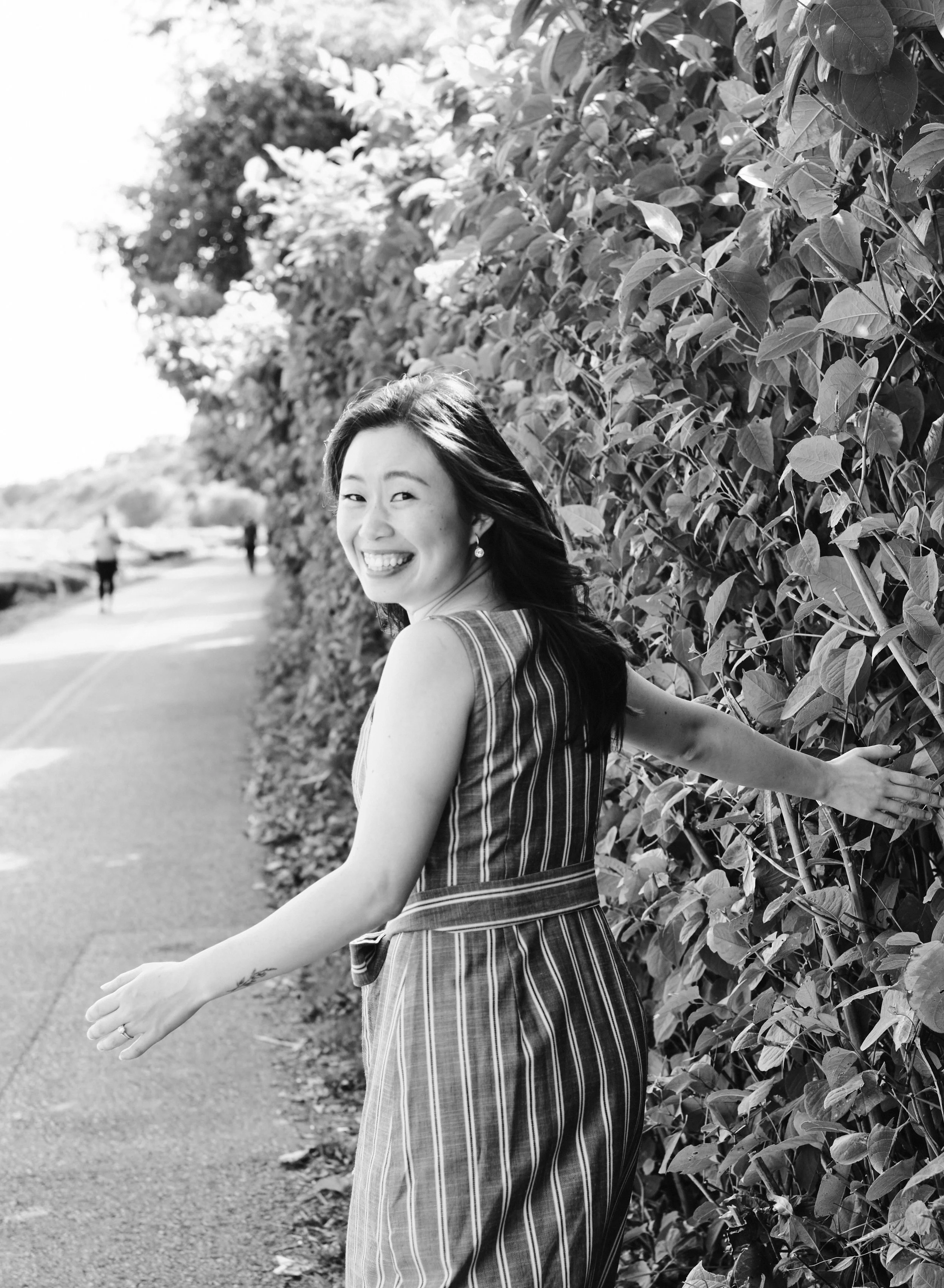 A woman smiling and holding onto a leafy hedge on a scenic outdoor path, in black and white.