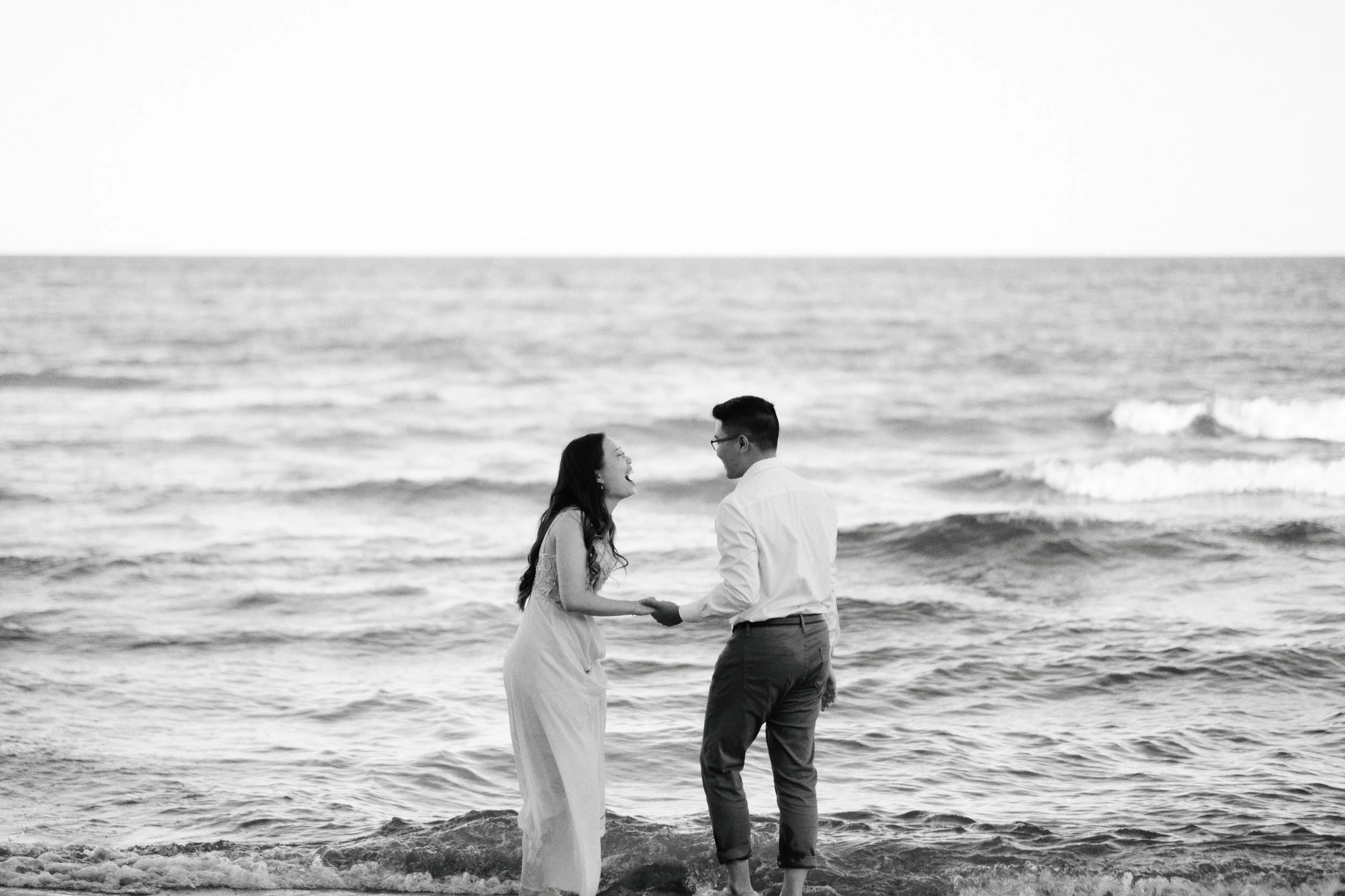A black and white photo of a couple standing on the beach holding hands, with the ocean waves in the background.