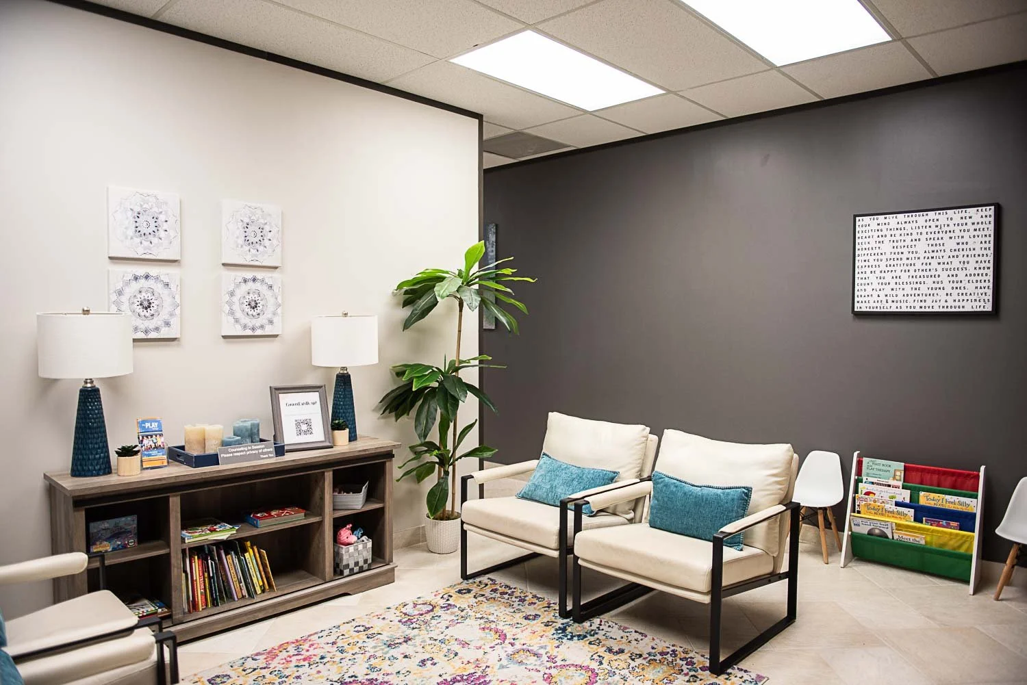 A cozy waiting room at Anchored by Light Counseling, PLLC, with two white armchairs, a bookshelf filled with books, and a side table with lamps, framed art, plants, and decorative items. A colorful rug covers the floor, with a word art on the dark gr