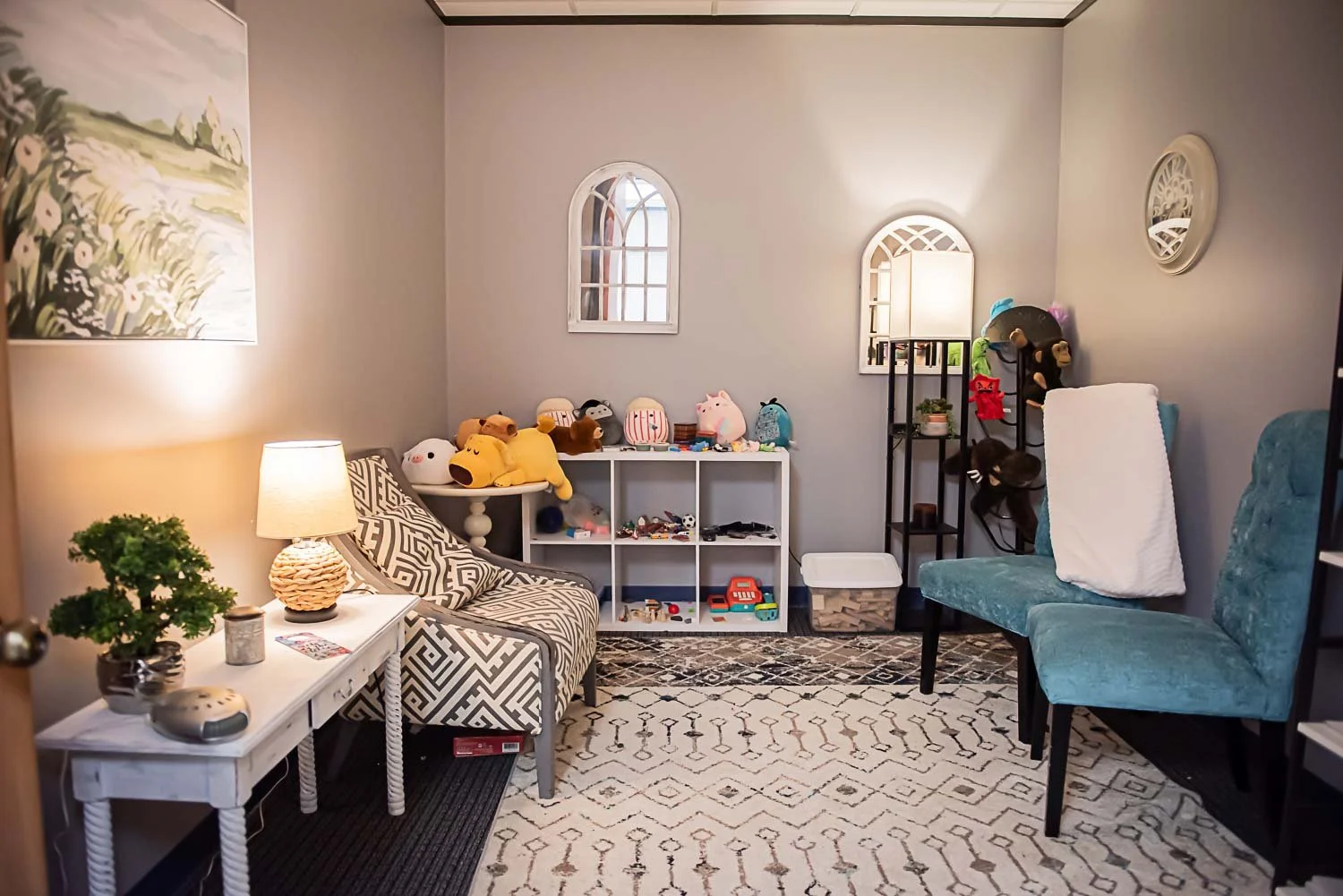 Private counseling office with gray walls, a patterned armchair with multicolored pillows, a white shelf with plush toys and small toys, a black metal shelving unit with stuffed animals, wallpaper on the side, a blue upholstered chair with a white bl