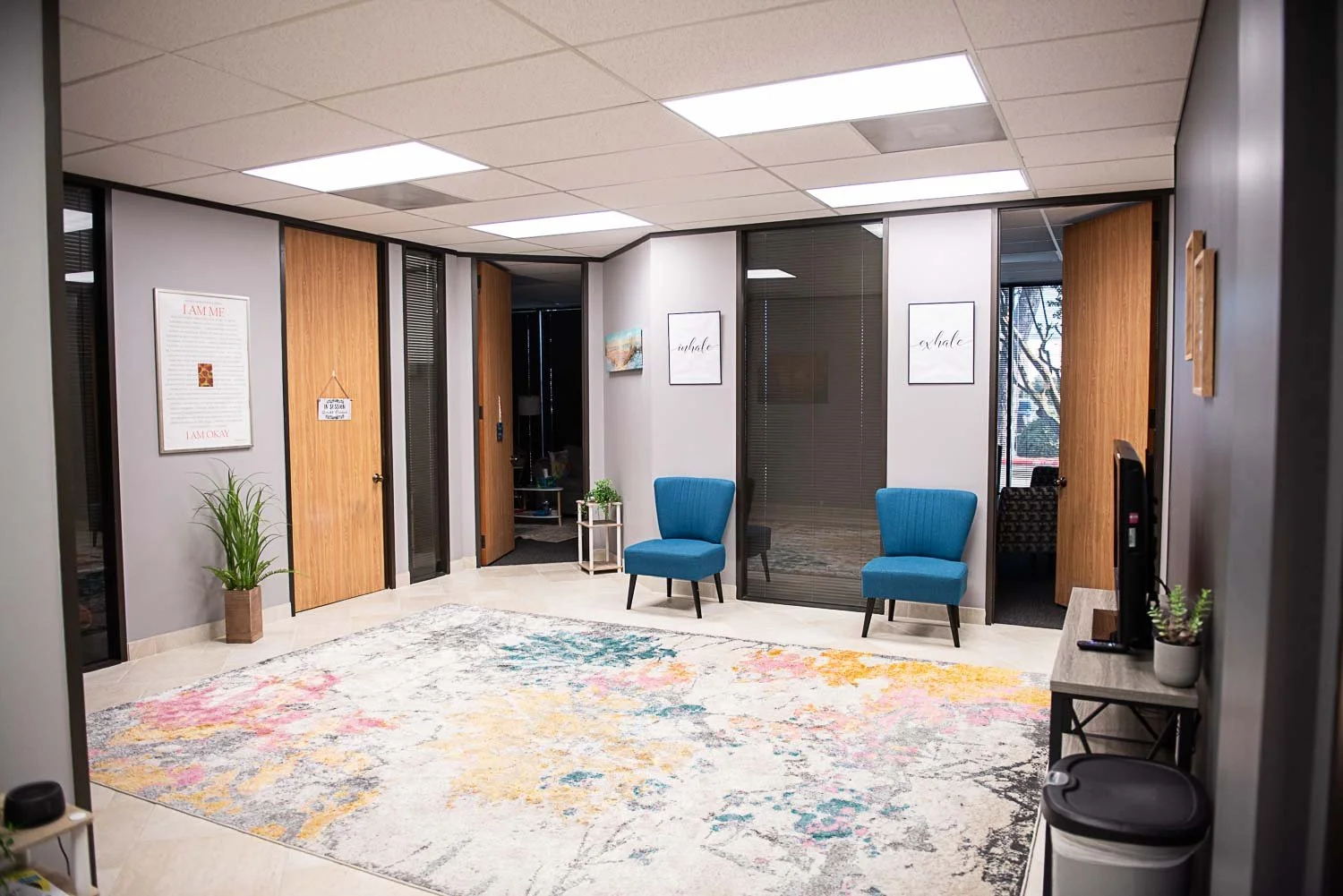 Office waiting area at Anchored by Light Counseling, PLLC, with two blue chairs, a colorful rug, potted plants, framed artwork on the walls, and closed doors leading to private offices.
