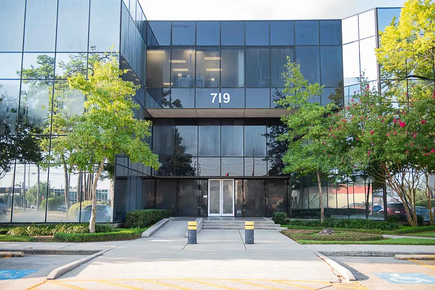 Modern glass office building with the number 719 on it, surrounded by greenery and trees, with a wheelchair accessible parking space in front.