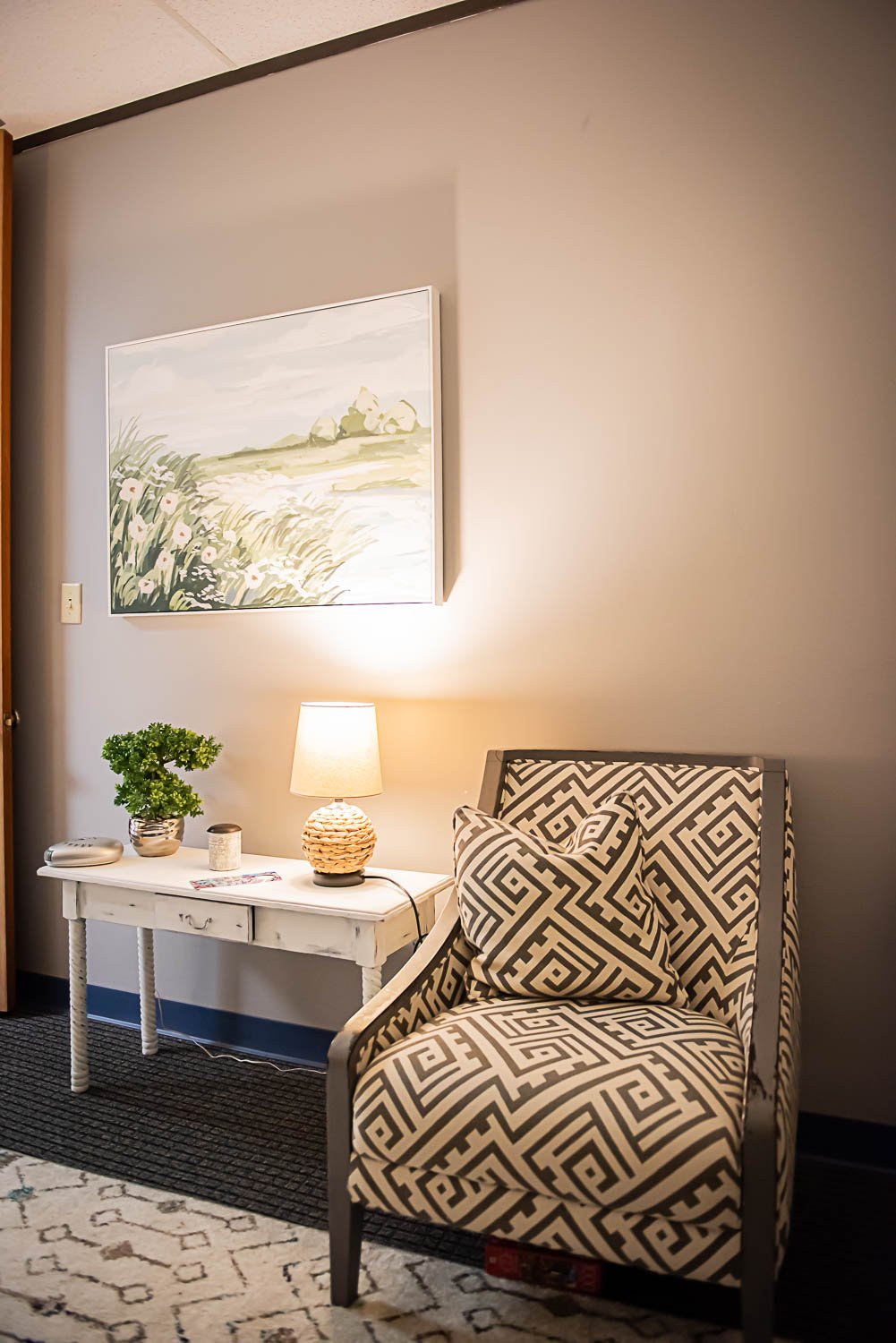 Private counseling office corner with patterned armchair, white side table with lamp, potted plant, and wall art of a landscape painting at Anchored by Light Counseling, PLLC.