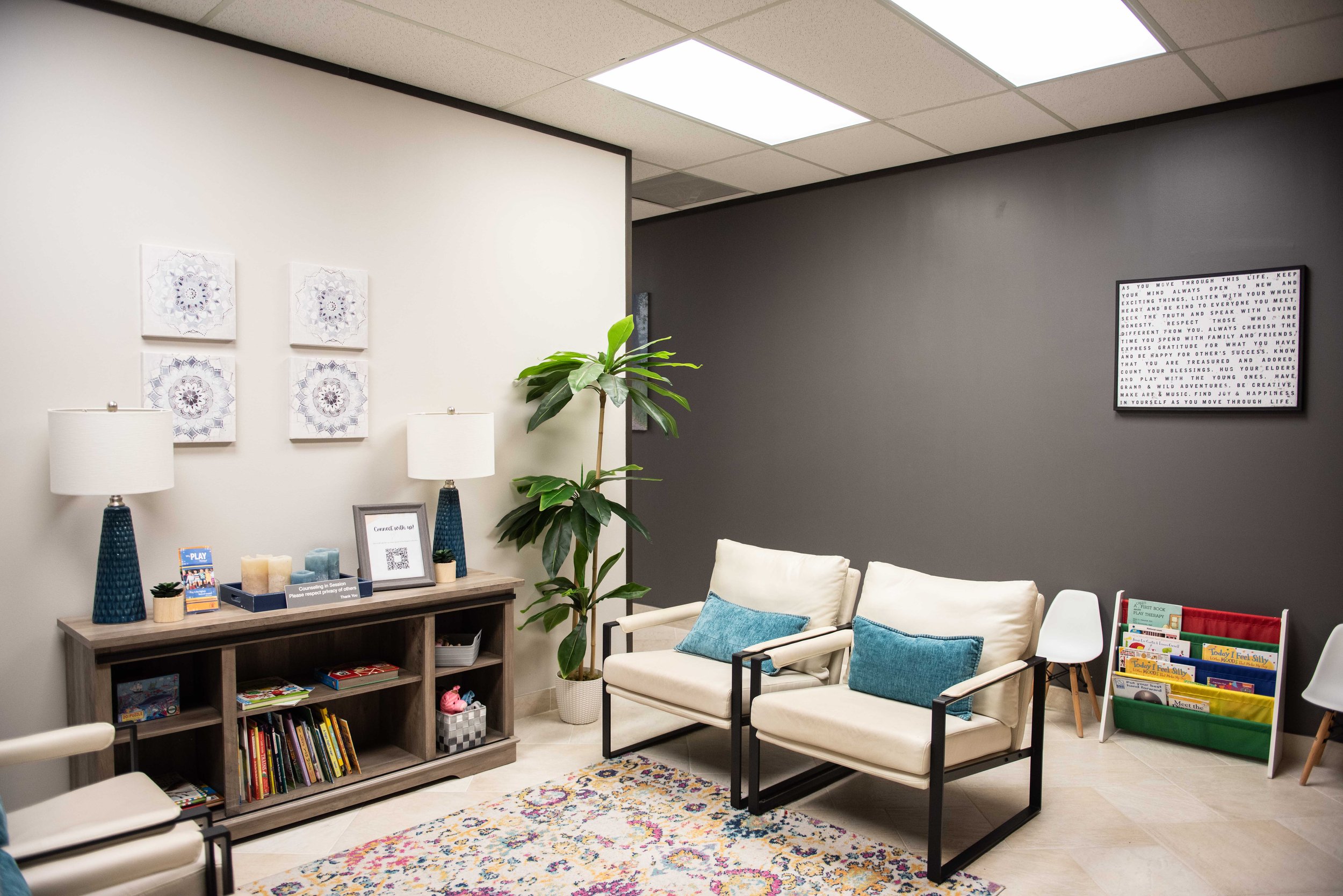 A cozy waiting room at Anchored by Light Counseling, PLLC, with two white armchairs, a bookshelf filled with books, and a side table with lamps, framed art, plants, and decorative items. A colorful rug covers the floor, with a word art on the dark gr