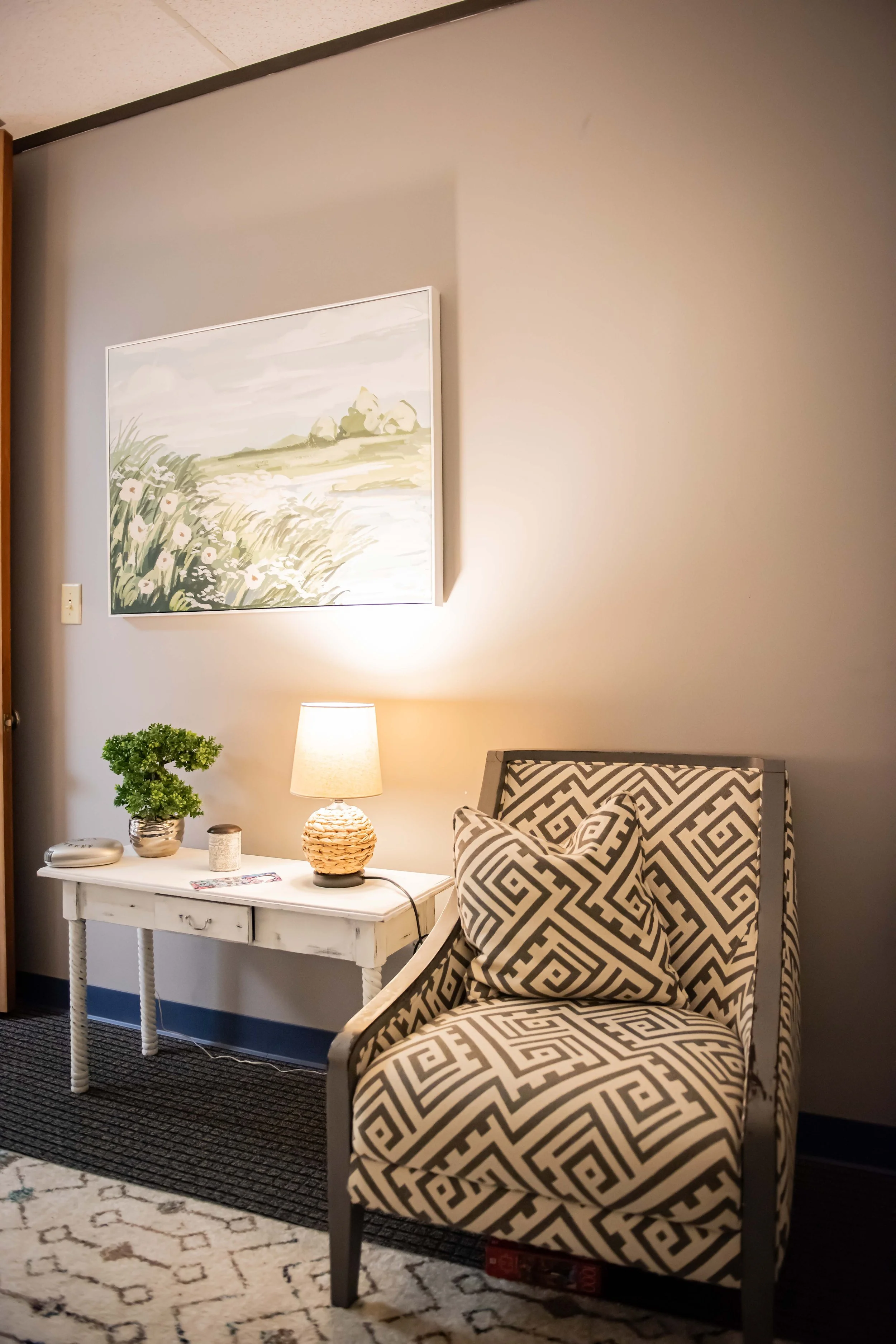 Private counseling office corner with patterned armchair, white side table with lamp, potted plant, and wall art of a landscape painting at Anchored by Light Counseling, PLLC.