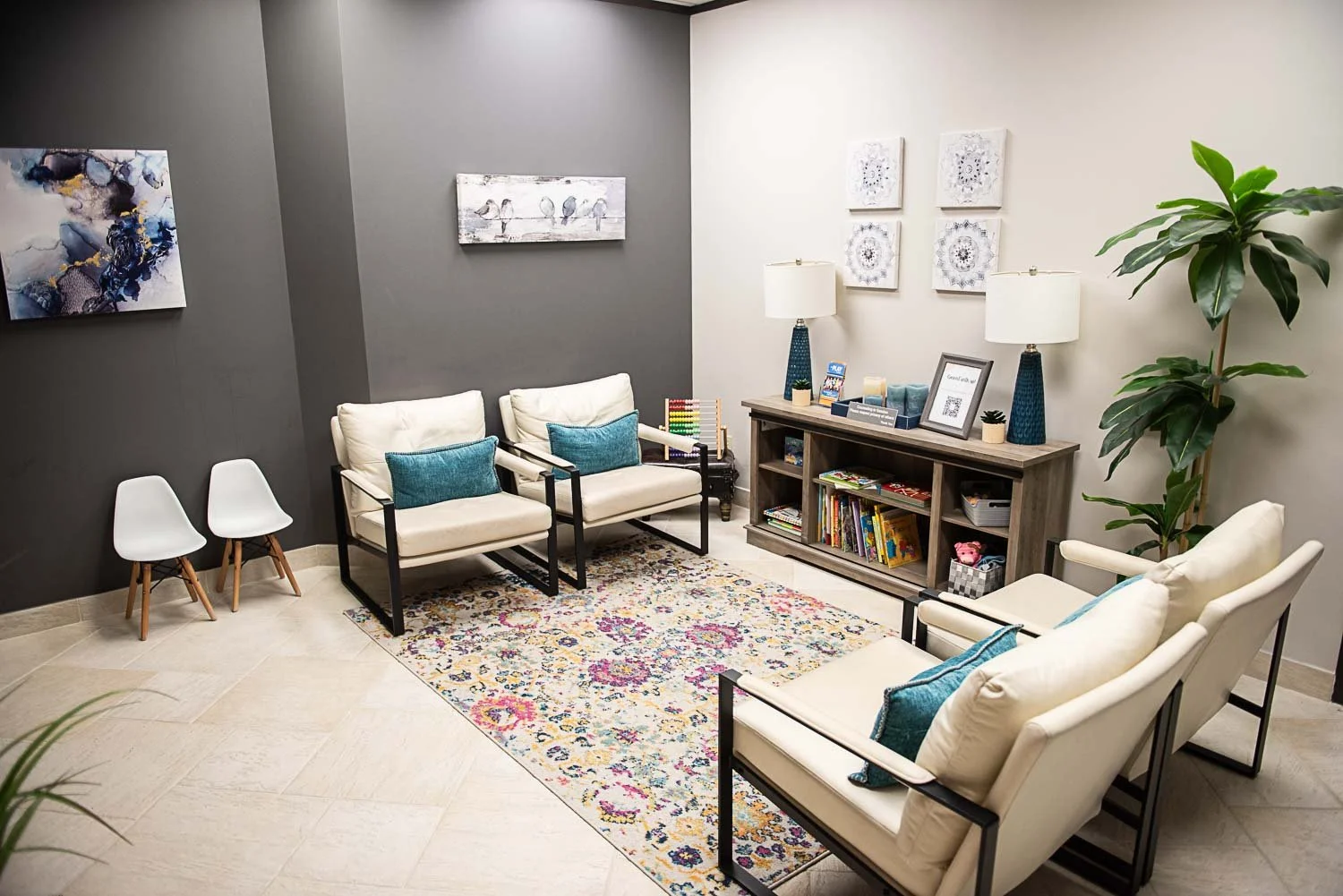 A waiting room at Anchored by Light Counseling, PLLC, with white chairs, a floral patterned rug, a dark gray accent wall, a bookshelf with books, and framed artwork, decorative lamps, a large plant, and toys.