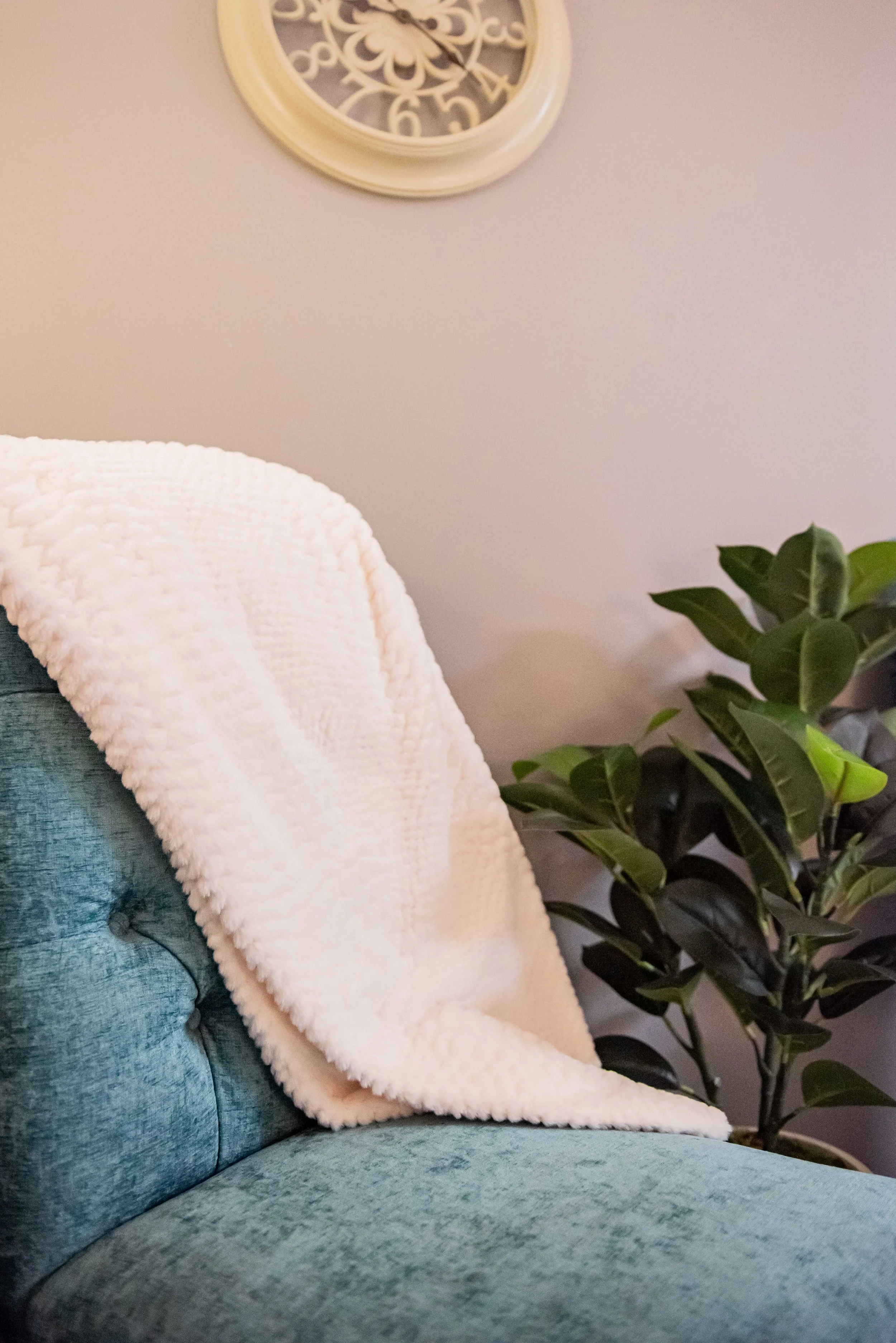 A light cream, textured blanket draped over the back of a teal armchair. A large leafy green plant next to the chair, and a round wall clock with a decorative face hanging above in a private counseling room at Anchored by Light Counseling, PLLC.