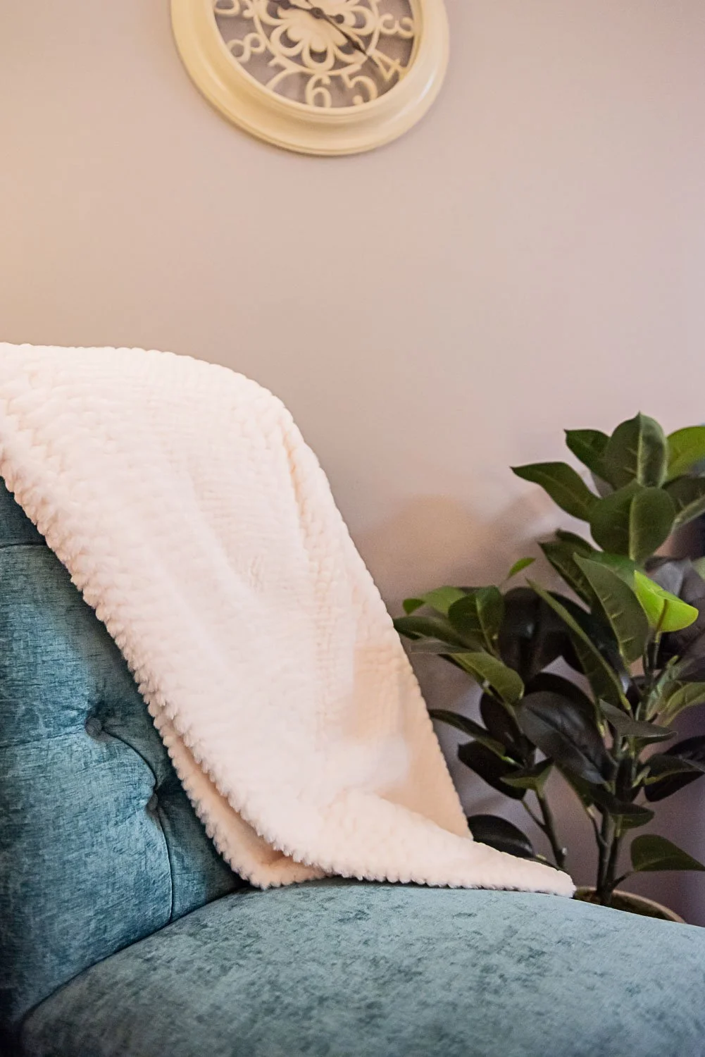 A light cream, textured blanket draped over the back of a teal armchair. A large leafy green plant next to the chair, and a round wall clock with a decorative face hanging above in a private counseling room at Anchored by Light Counseling, PLLC.