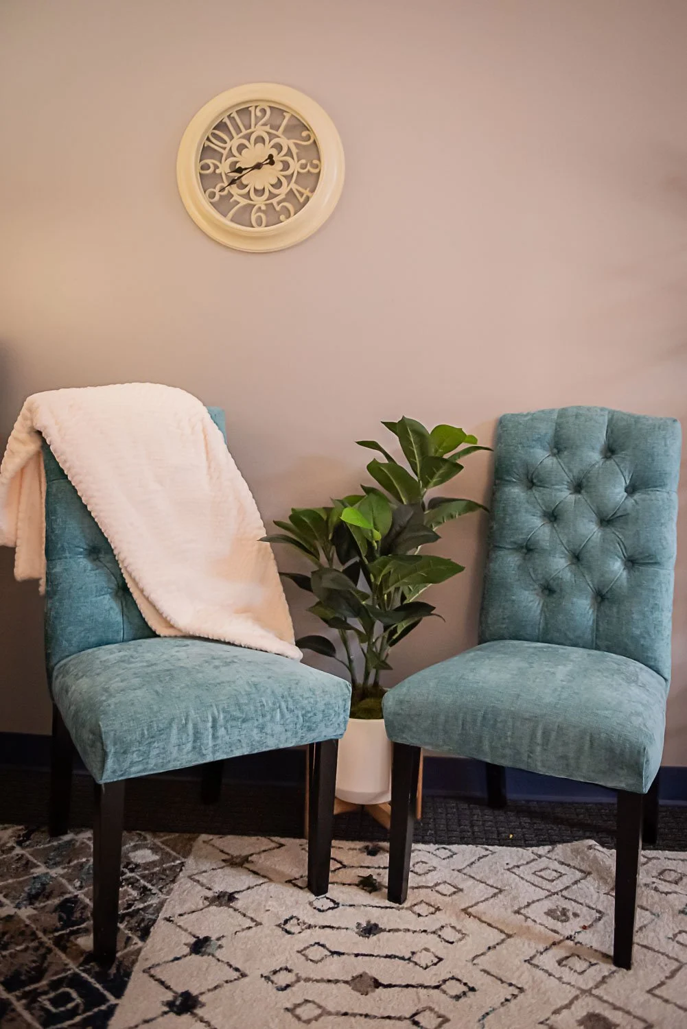 Two teal upholstered chairs with black legs, a potted plant between them, a white blanket draped over one chair, a wall clock above, and a patterned rug on the floor in private counseling office at Anchored by Light Counseling, PLLC.