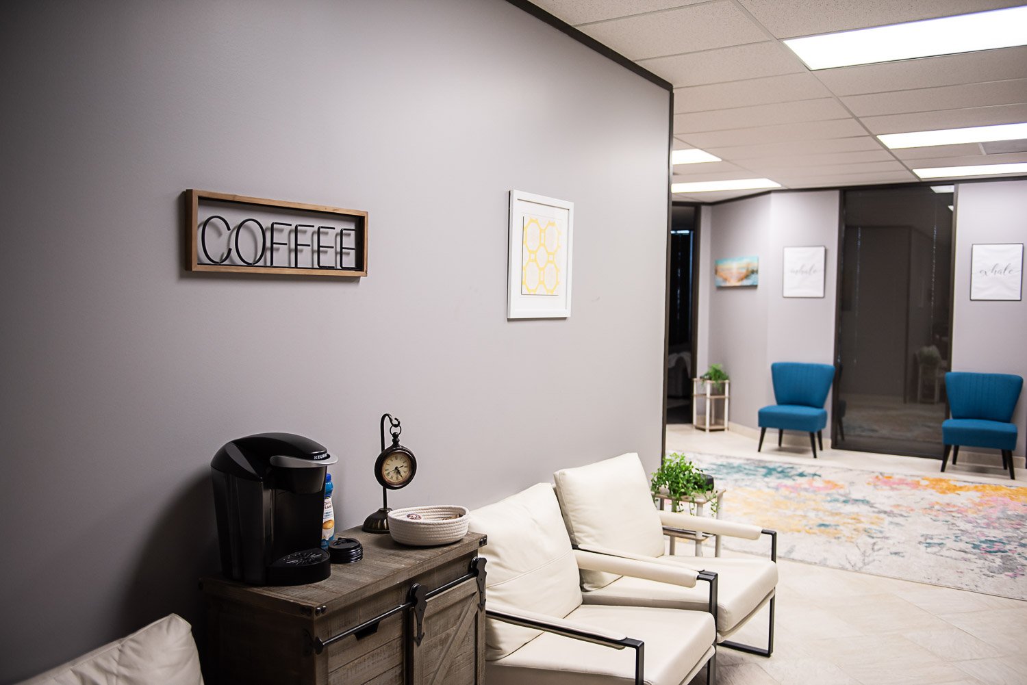 Lobby area at Anchored by Light Counseling, PLLC, with a coffee station, white armchairs, a colorful rug, and blue chairs, with artwork on the walls and a window behind the chairs.