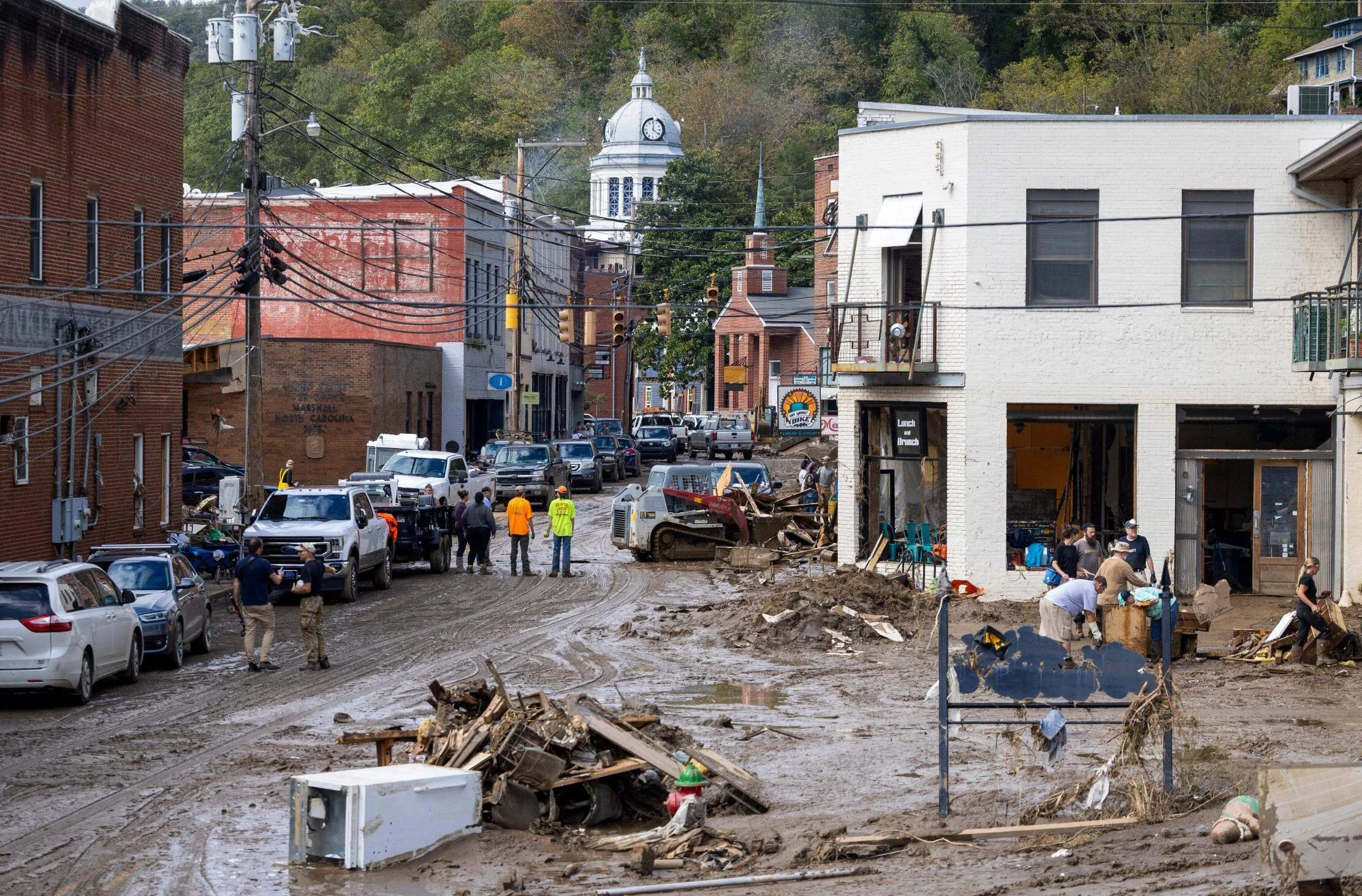 A muddy street in a small town with several people, vehicles, and damaged buildings, likely after a flood or disaster, with construction equipment and debris on the ground.
