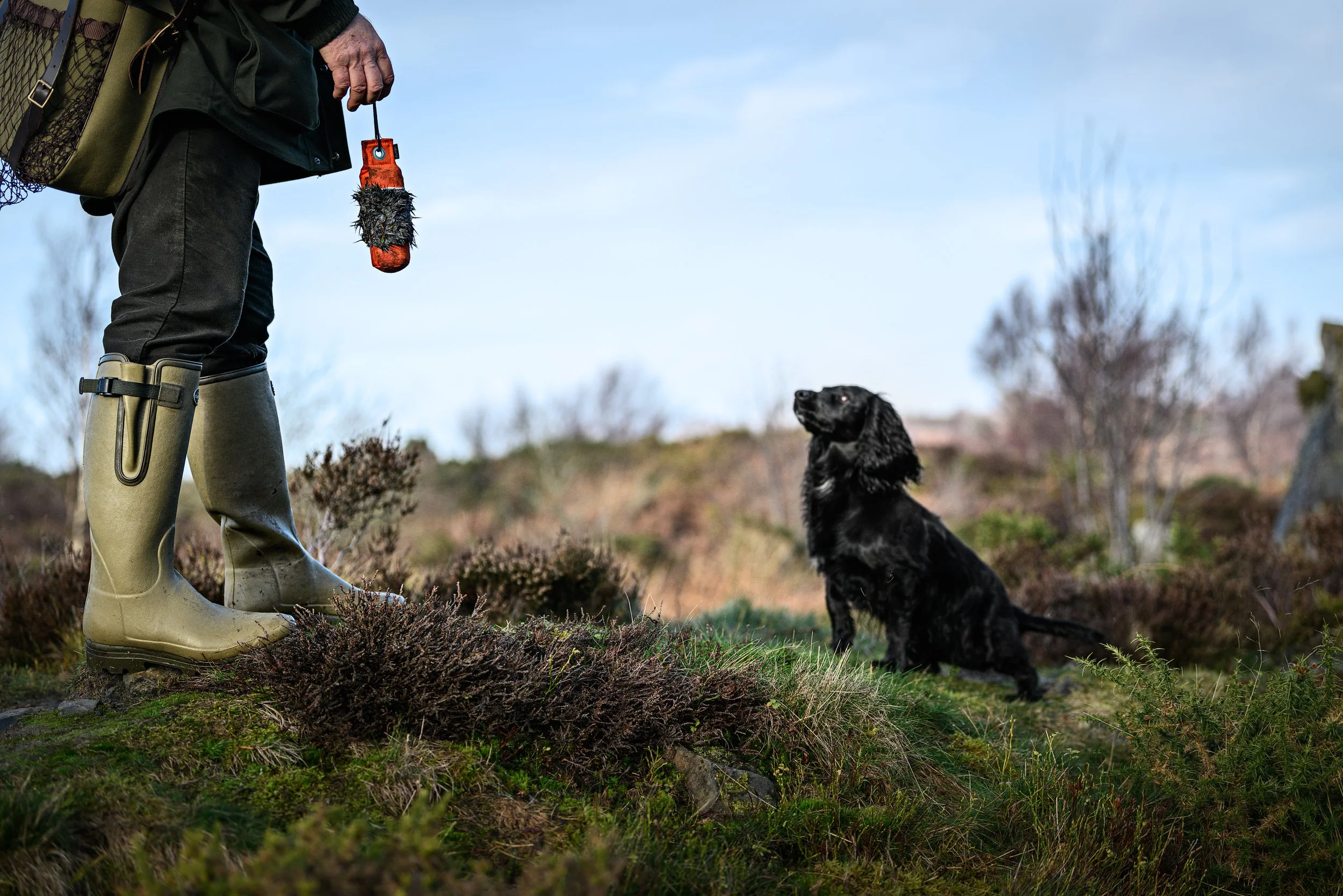 A person standing in a natural outdoor setting, dressed in rubber boots and outdoor clothing, holding a game bird launcher, with a black spaniel sitting attentively nearby.