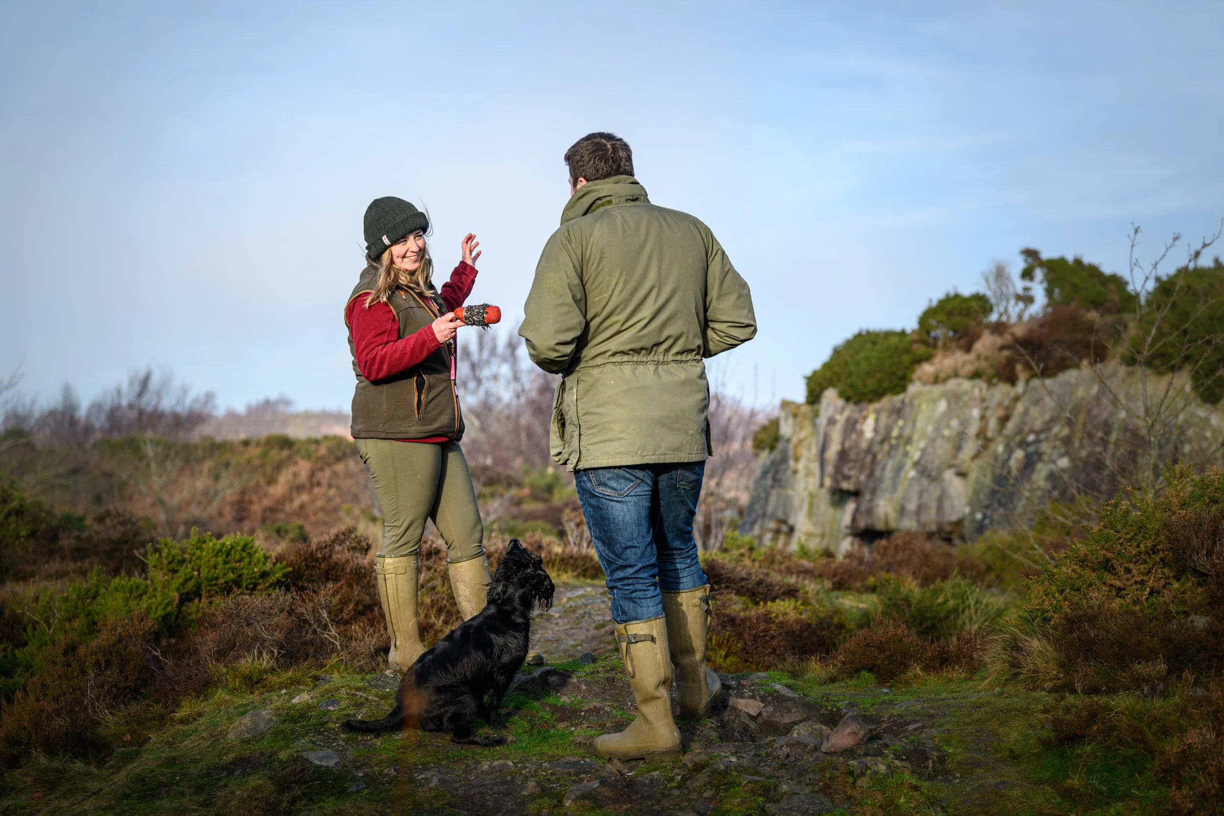 A woman and man stand outdoors in a natural landscape, engaging in a conversation with a black dog nearby. The woman is wearing a black beanie, red and brown jacket, khaki pants, and rubber boots, holding a small stick or toy. The man is dressed in a green jacket, blue jeans, and rubber boots. They are surrounded by shrubs and rocky terrain under a blue sky.
