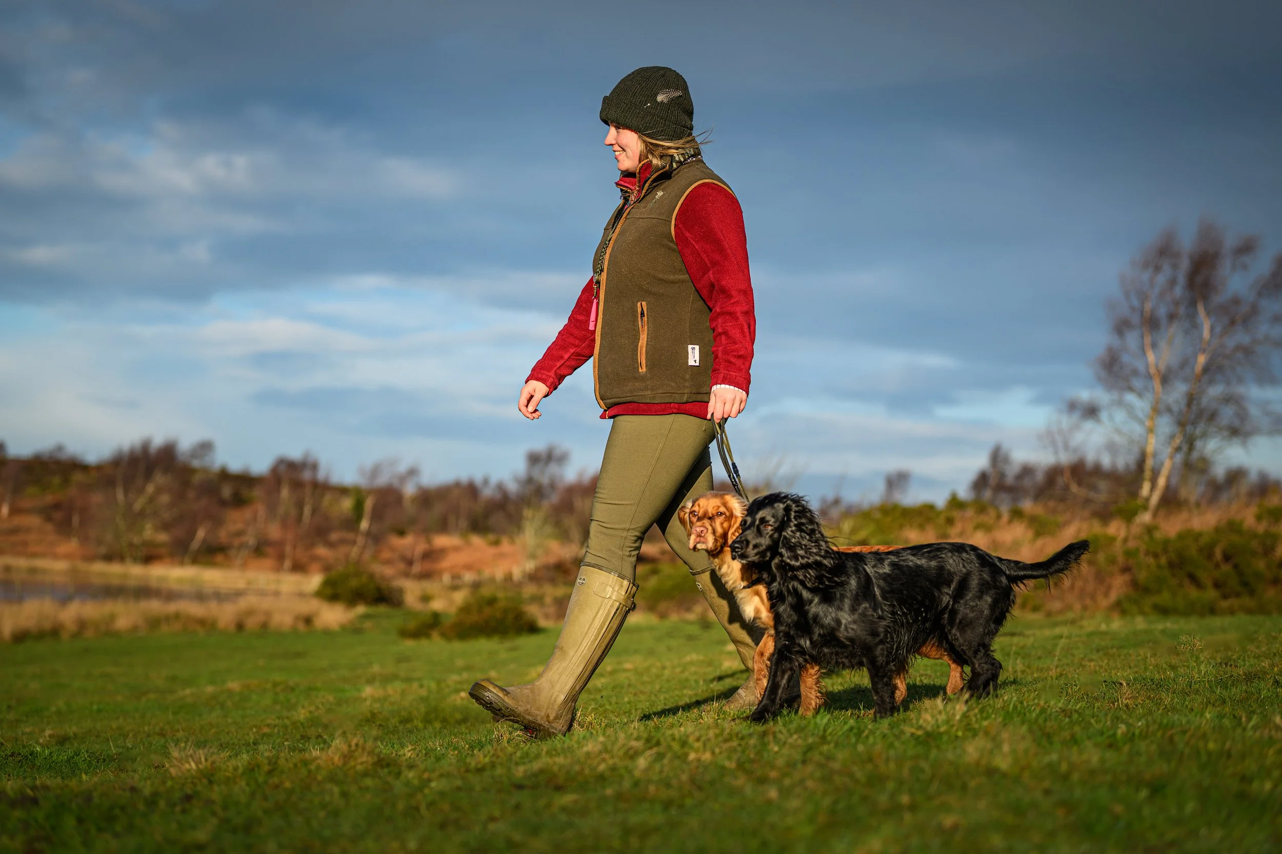 Woman walking two dogs on a grassy field under a cloudy sky.
